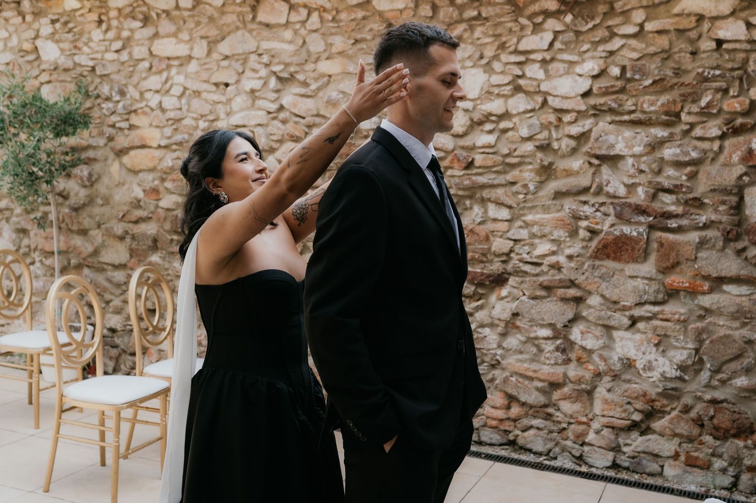 A bride in a black gown touches a groom's face during a first look moment against a rustic stone wall backdrop.