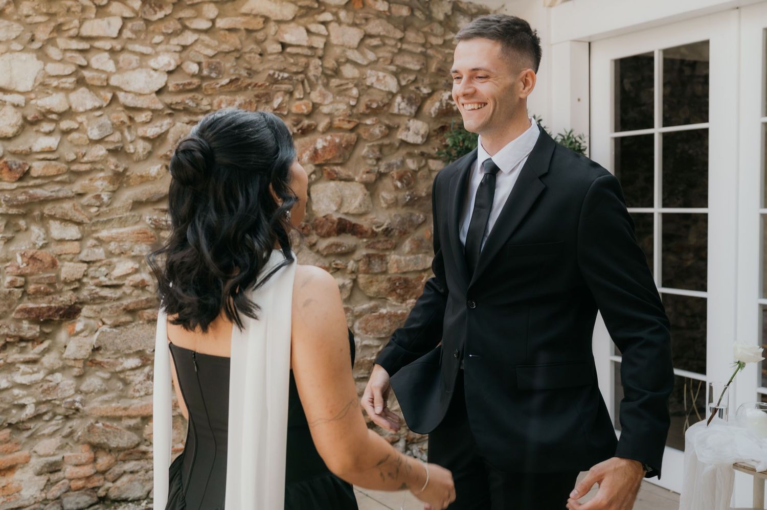 A smiling groom in a black suit holds hands with a bride in a black and white dress against a stone wall.