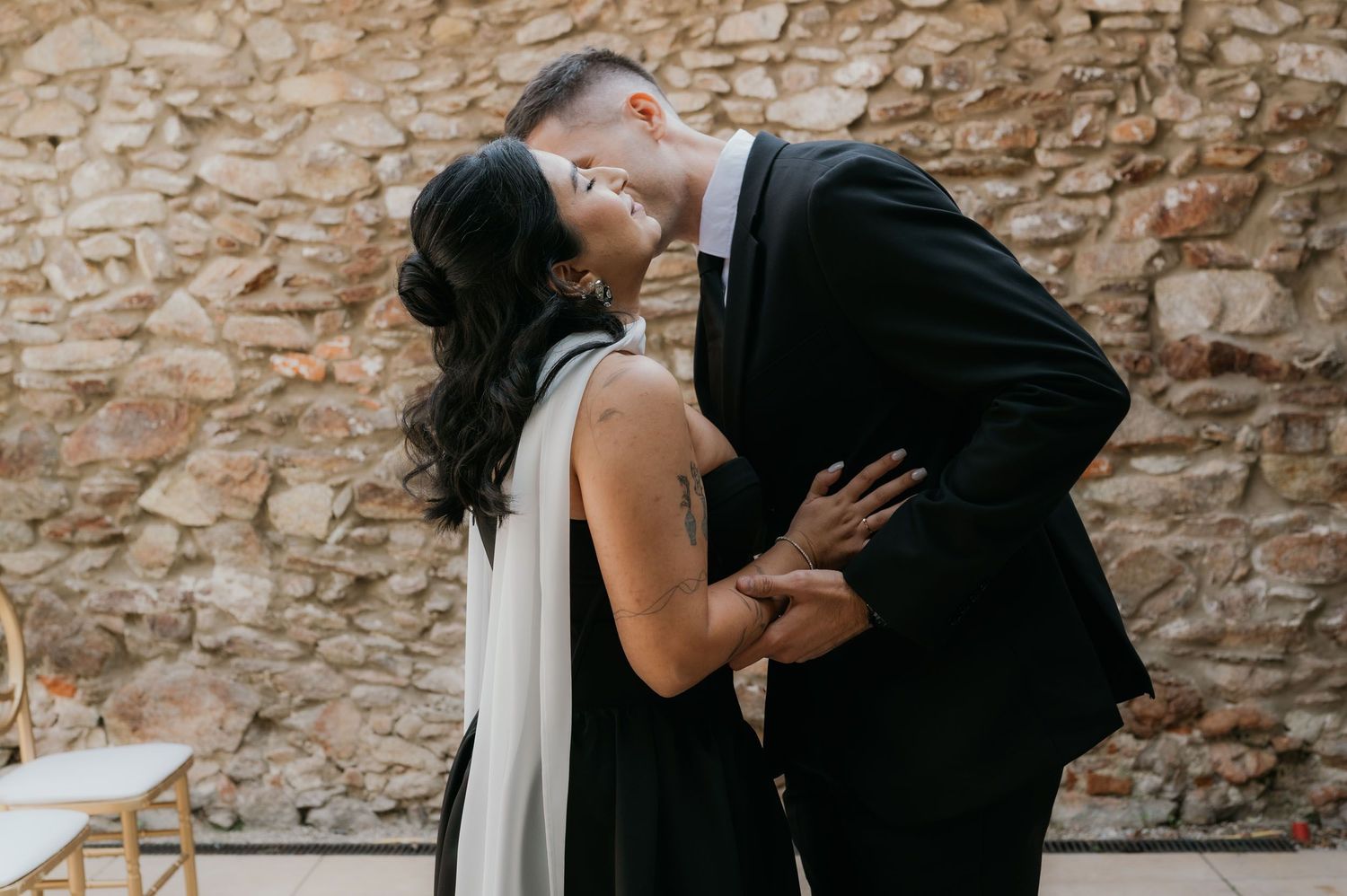 Couple sharing a romantic kiss in elegant black attire against a rustic stone wall backdrop.