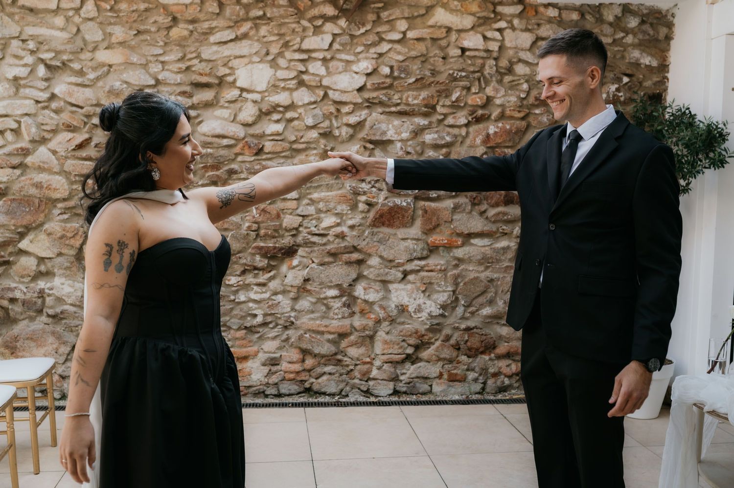 Couple in black attire touching hands in front of a rustic stone wall at an elegant indoor venue.