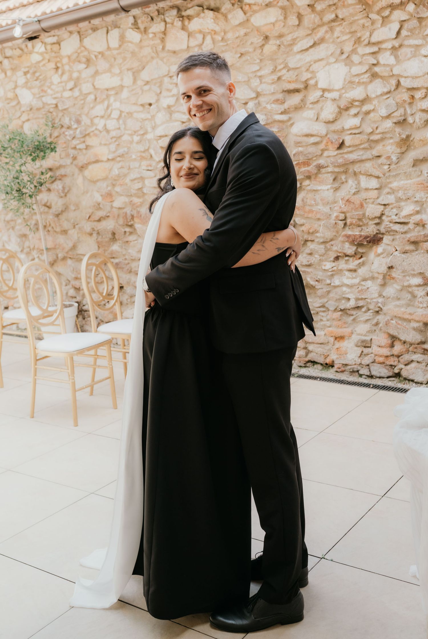 Couple embracing at outdoor wedding venue, woman in black and white gown, man in black suit, stone wall backdrop.