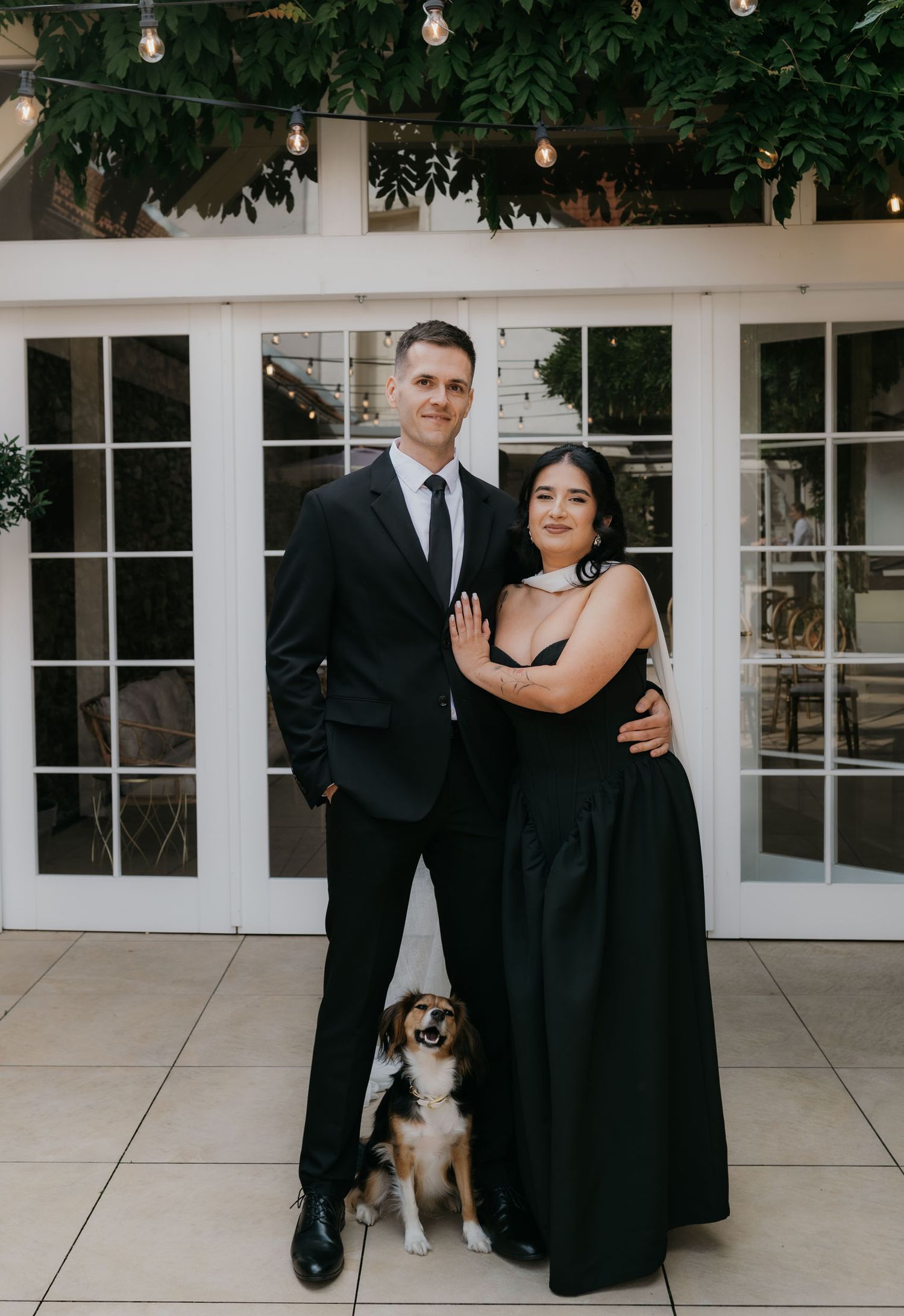 Couple in elegant black formal attire posing with a small beagle dog on a patio with string lights.