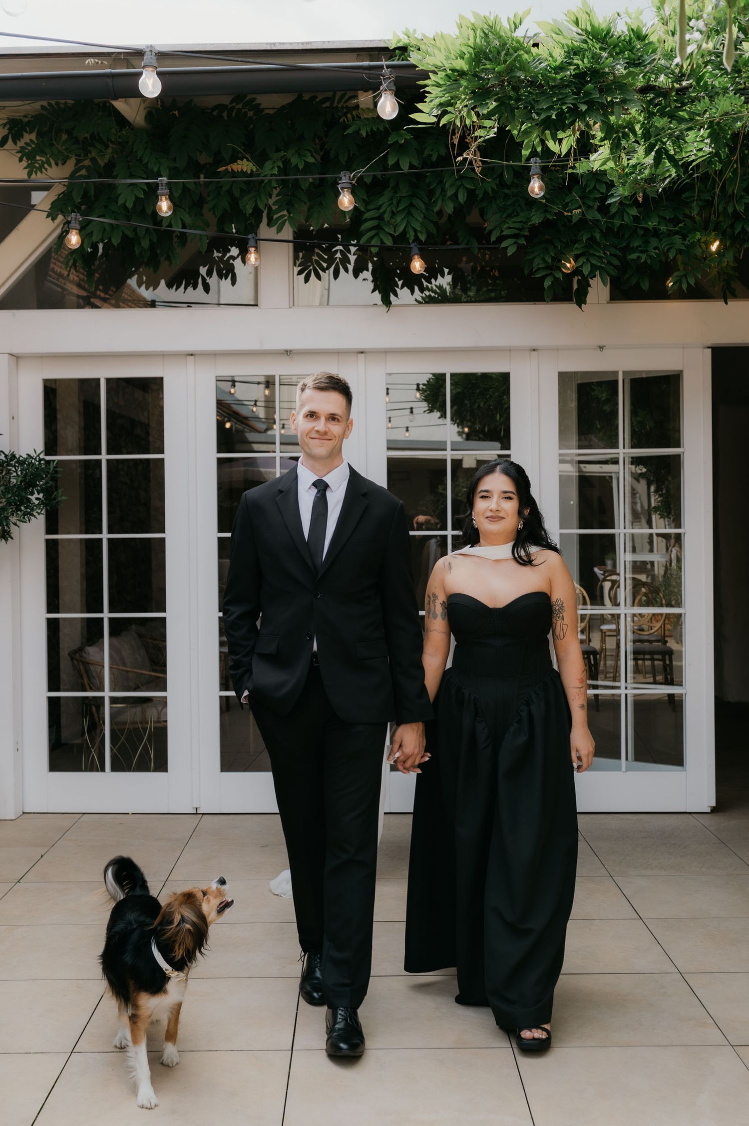 Couple in matching black formal attire holding hands with their dog at an elegant outdoor venue with string lights.