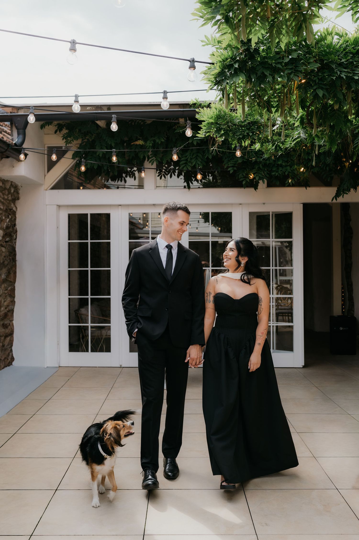 Couple in matching black formal attire hold hands with small dog on patio with string lights and lush greenery.