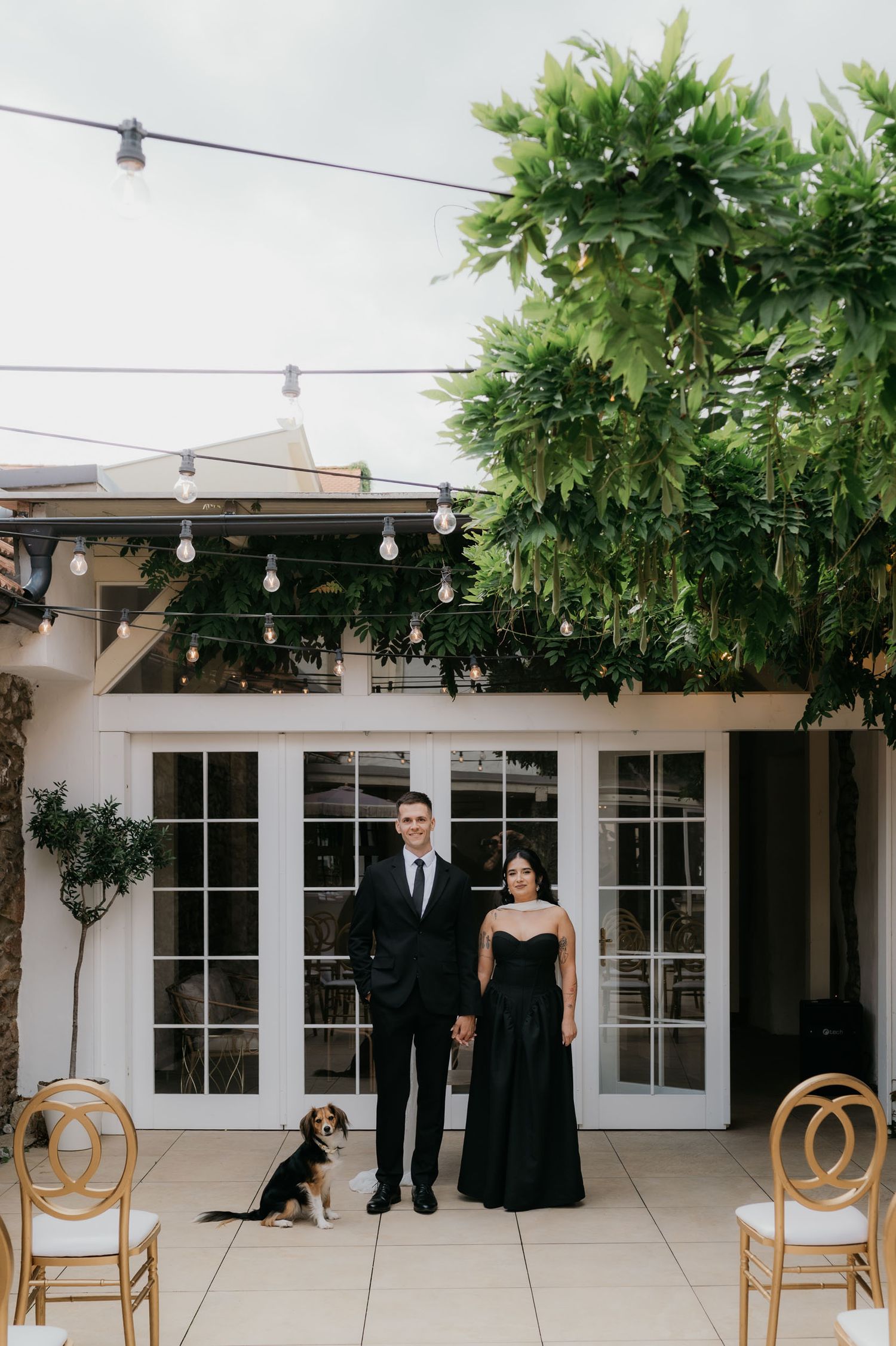 Couple in matching black formal attire stand with their beagle in a white courtyard with string lights and French doors.