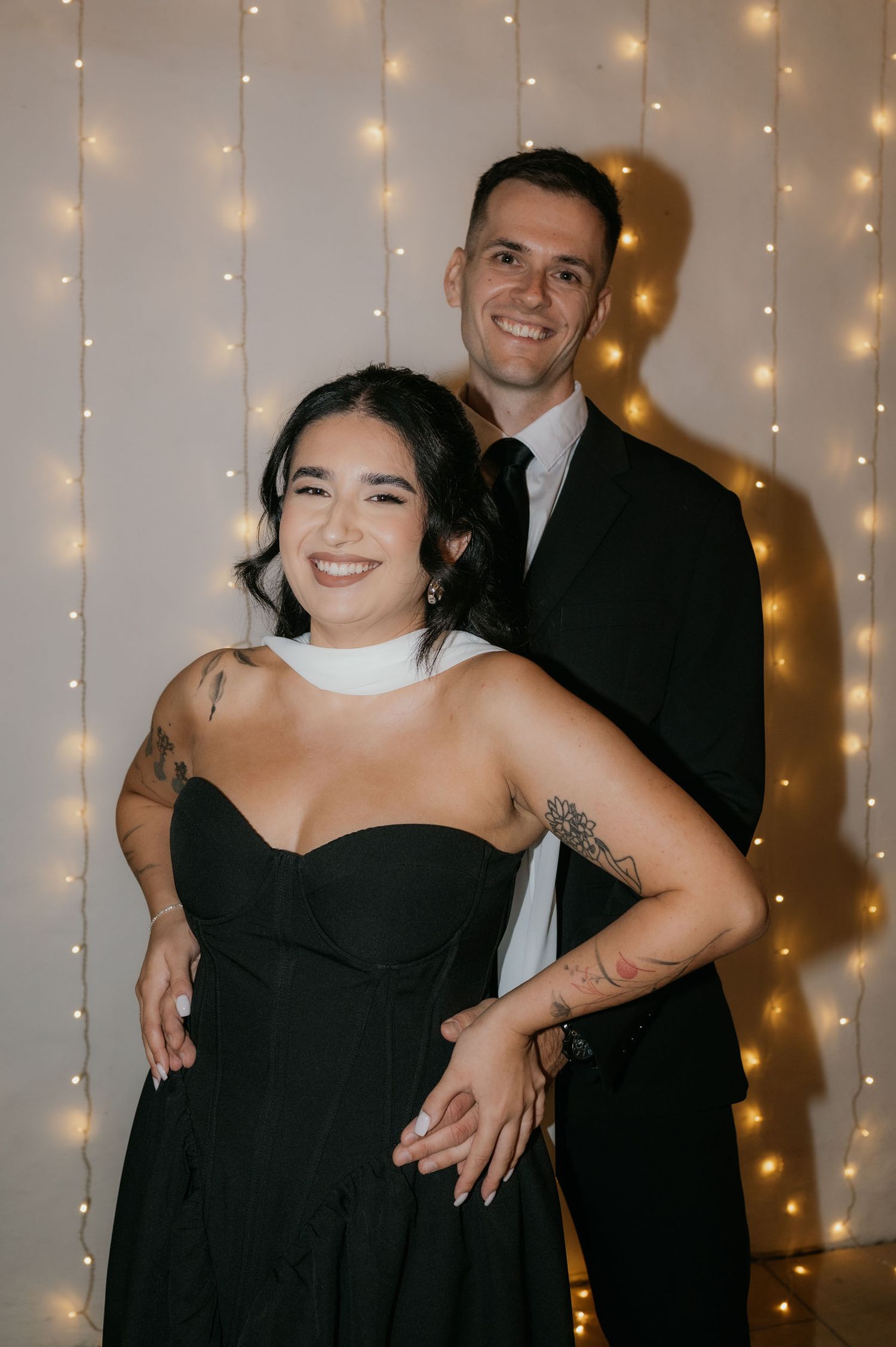 Couple posing together in black formal wear against a twinkling fairy light backdrop at a wedding reception.