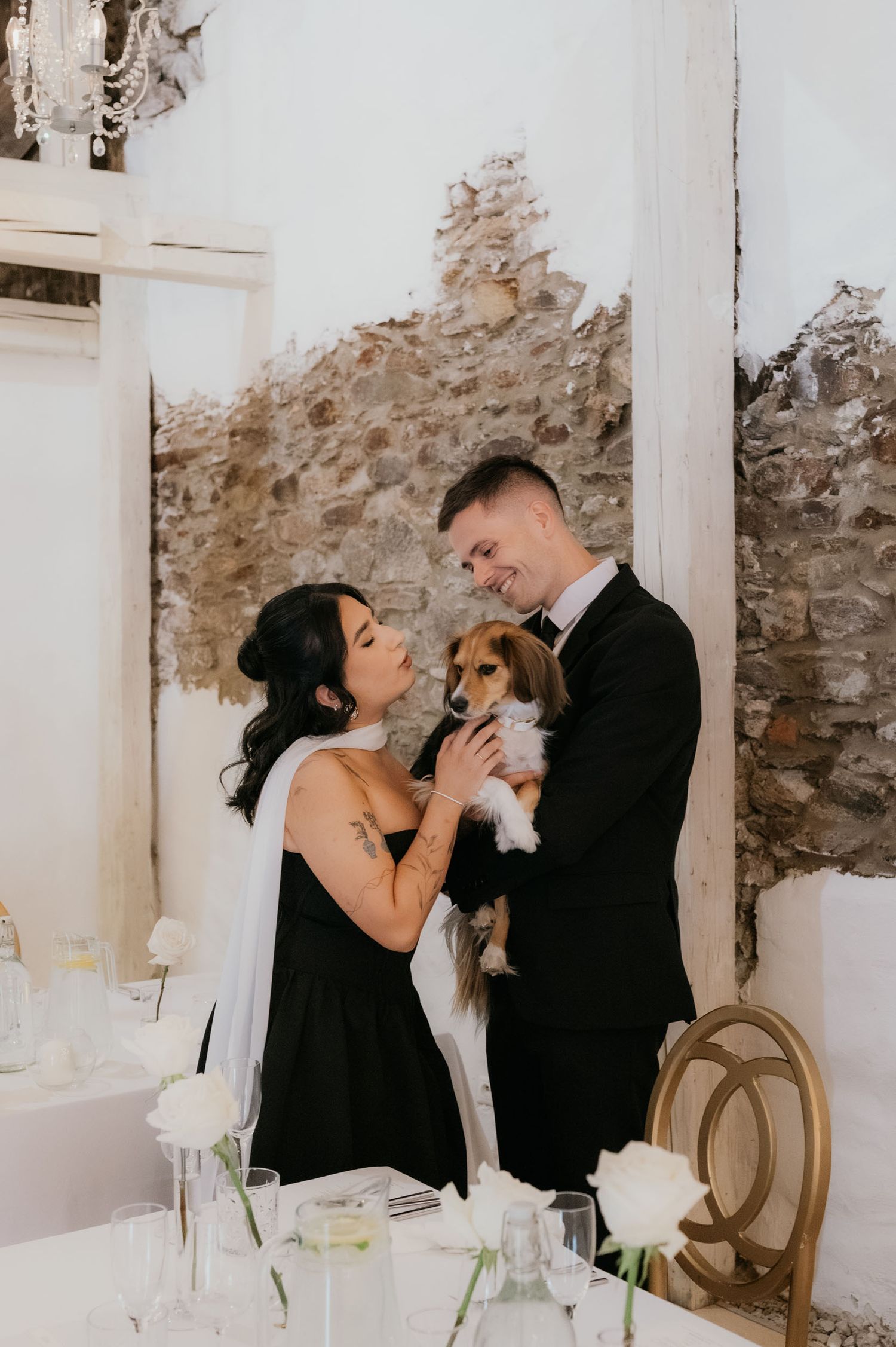 Bride and groom in black attire holding a small dog at elegant wedding reception with stone wall backdrop.