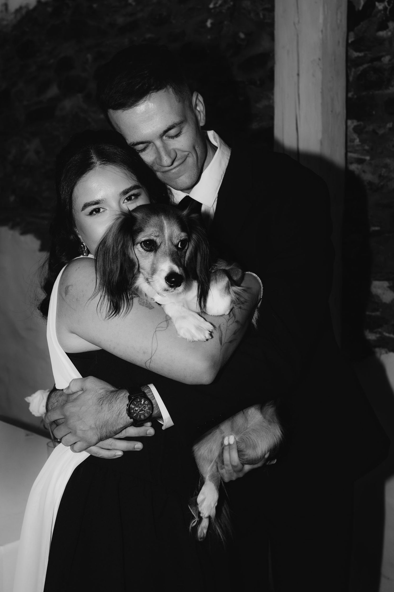 Black and white photo of a couple in formal wedding attire embracing while holding their small dog.