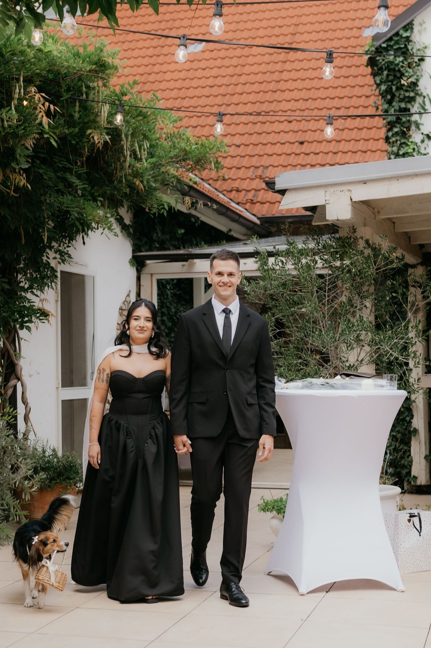 A couple in matching black formal attire pose together at an outdoor wedding venue with string lights.