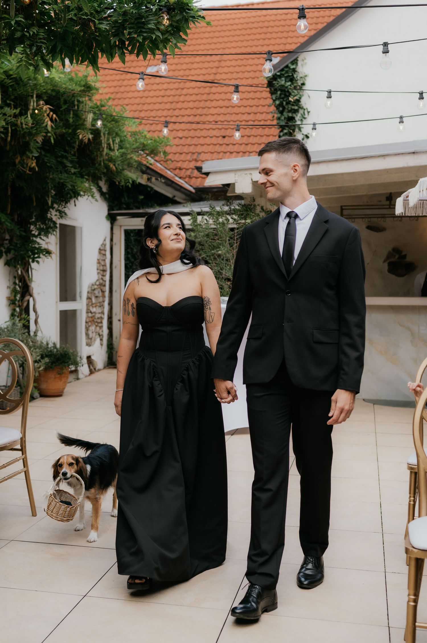 Couple in matching black wedding attire holding hands with a beagle dog in a floral wreath at an outdoor venue.