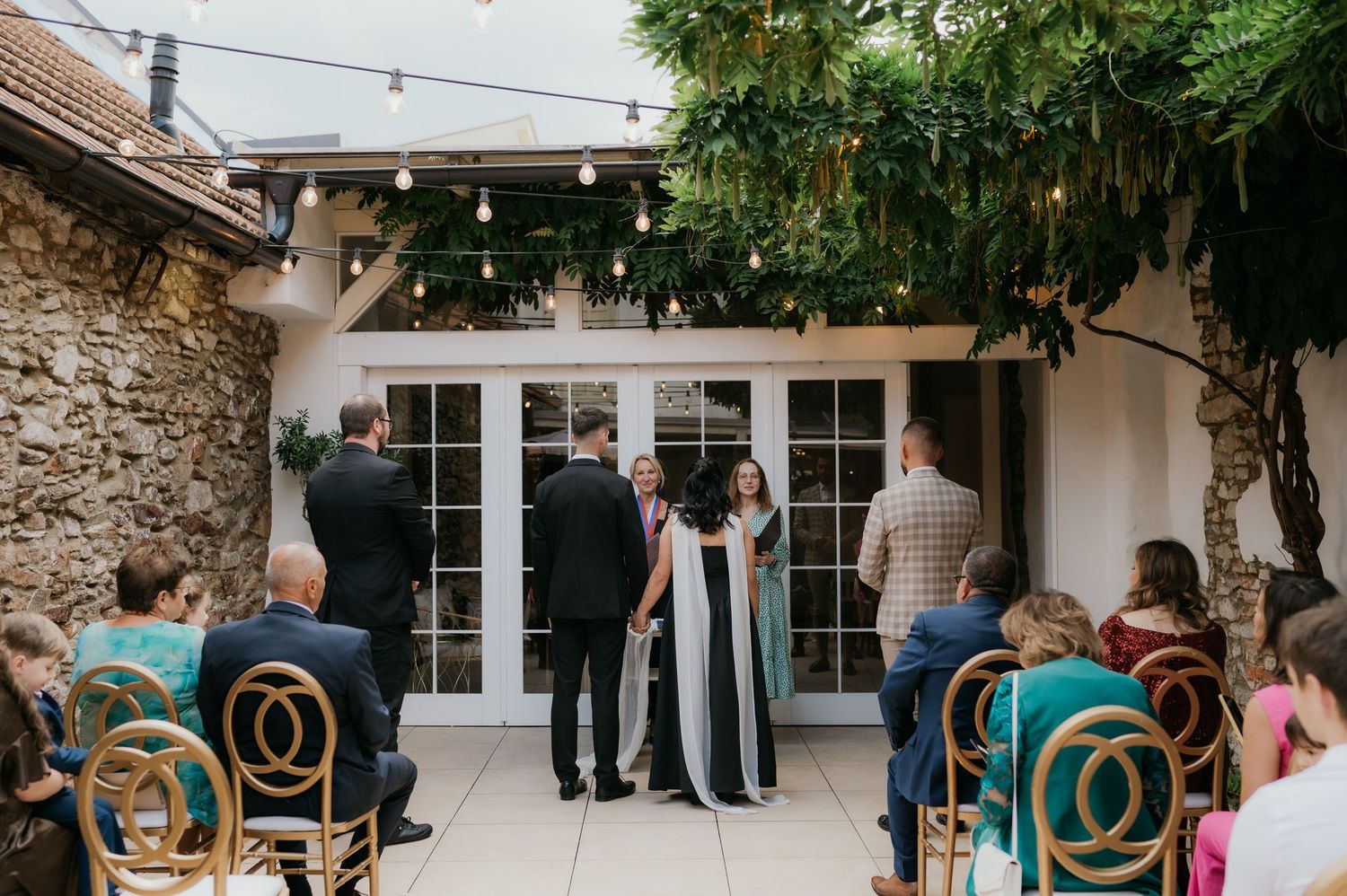 Guests seated at an outdoor wedding ceremony in a rustic courtyard with string lights and lush greenery.