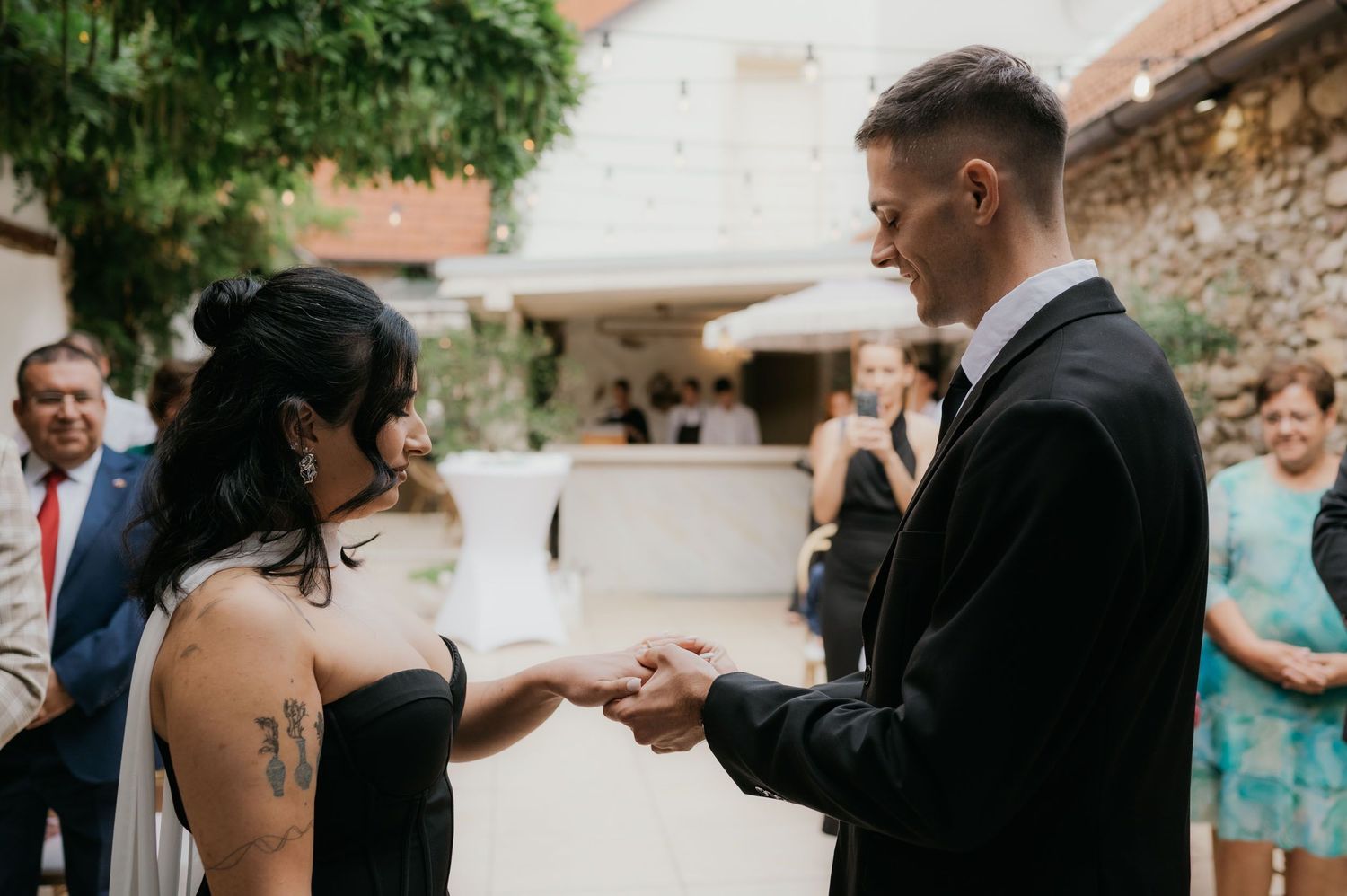 Bride and groom exchanging rings during outdoor wedding ceremony with guests watching in background.