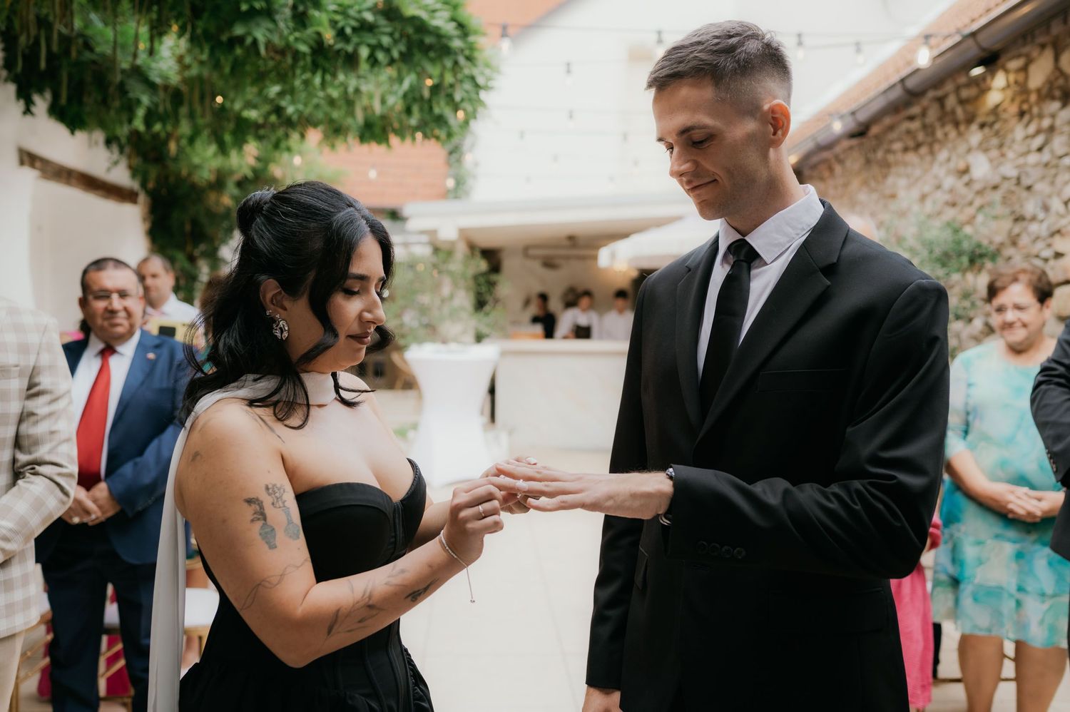 Tattooed bride in black dress placing wedding ring on groom's finger during outdoor ceremony.