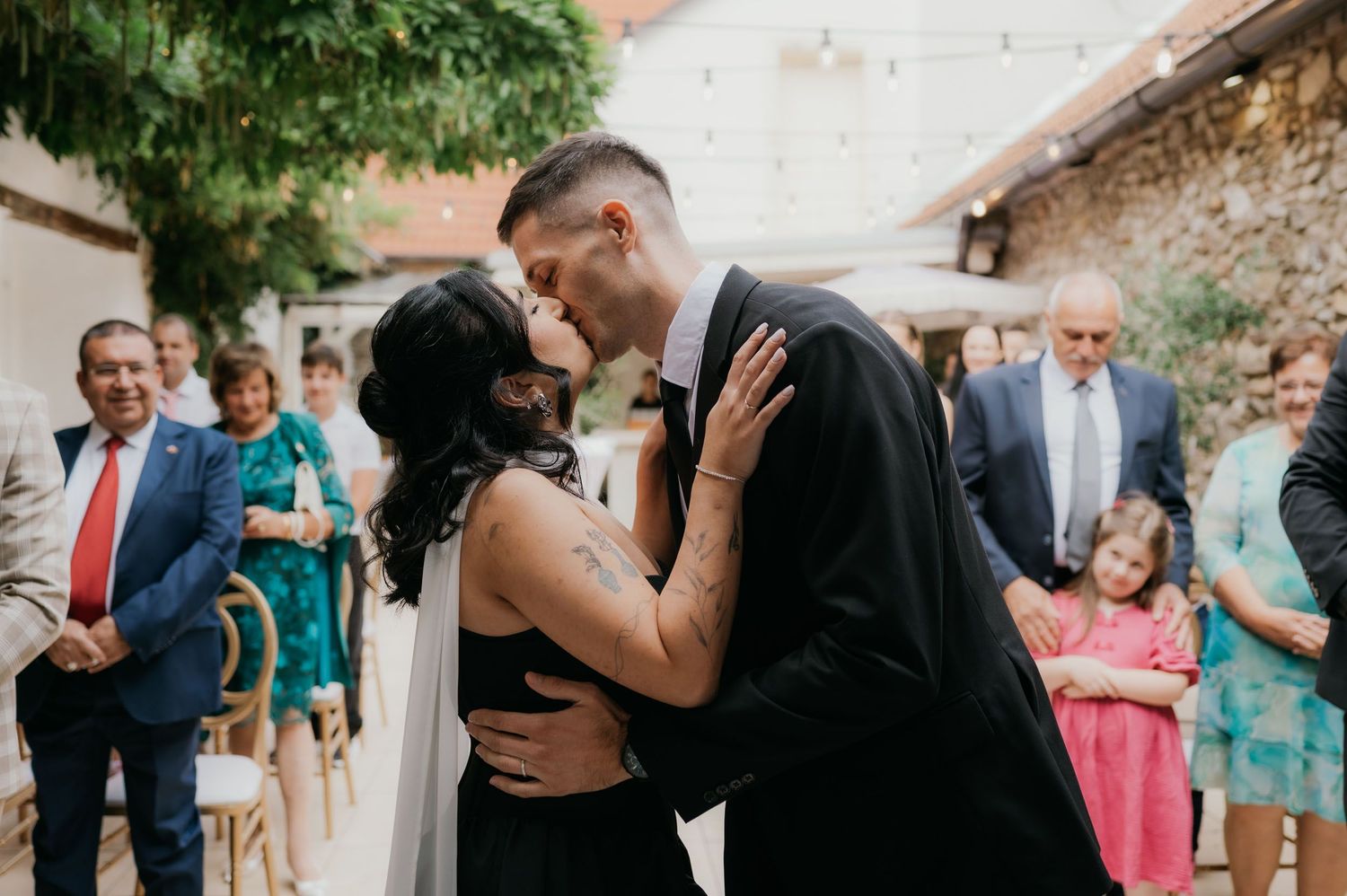 Couple sharing first kiss at outdoor wedding ceremony with guests watching in background.