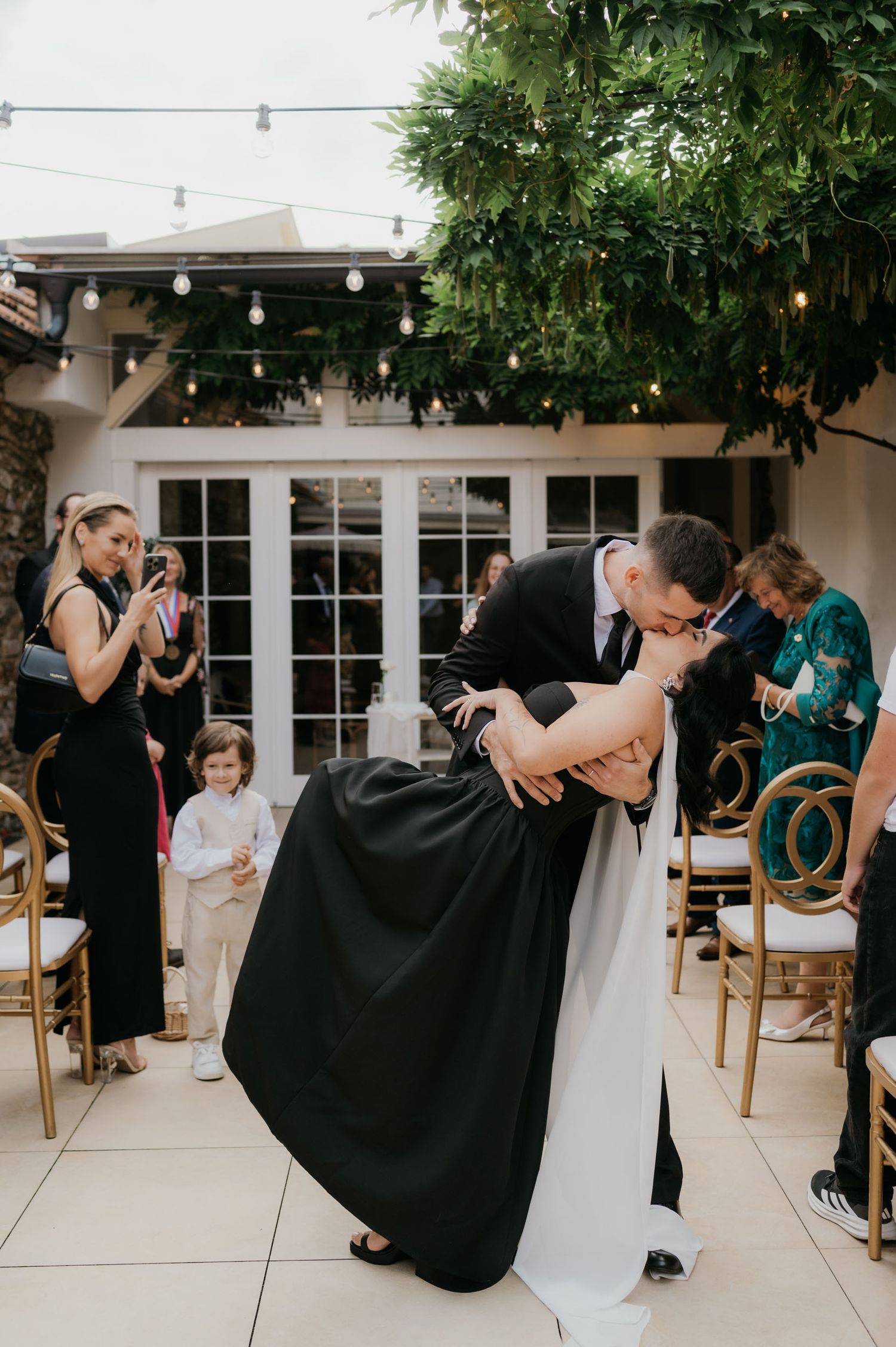 A groom dips and kisses his bride in a black and white wedding dress during their ceremony at an elegant outdoor venue.