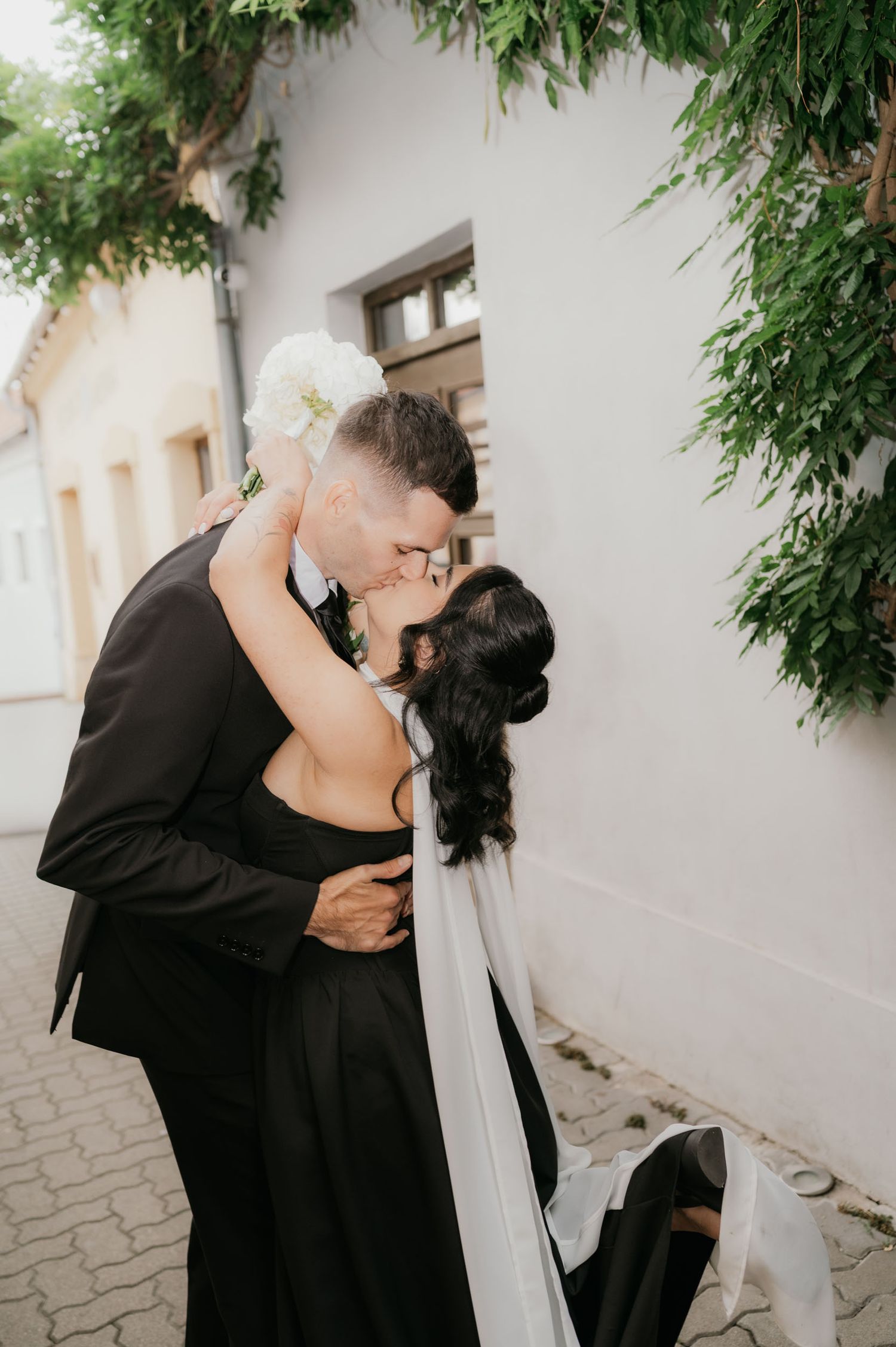 A bride and groom share a romantic kiss outdoors, dressed in elegant black wedding attire with a flowing veil.