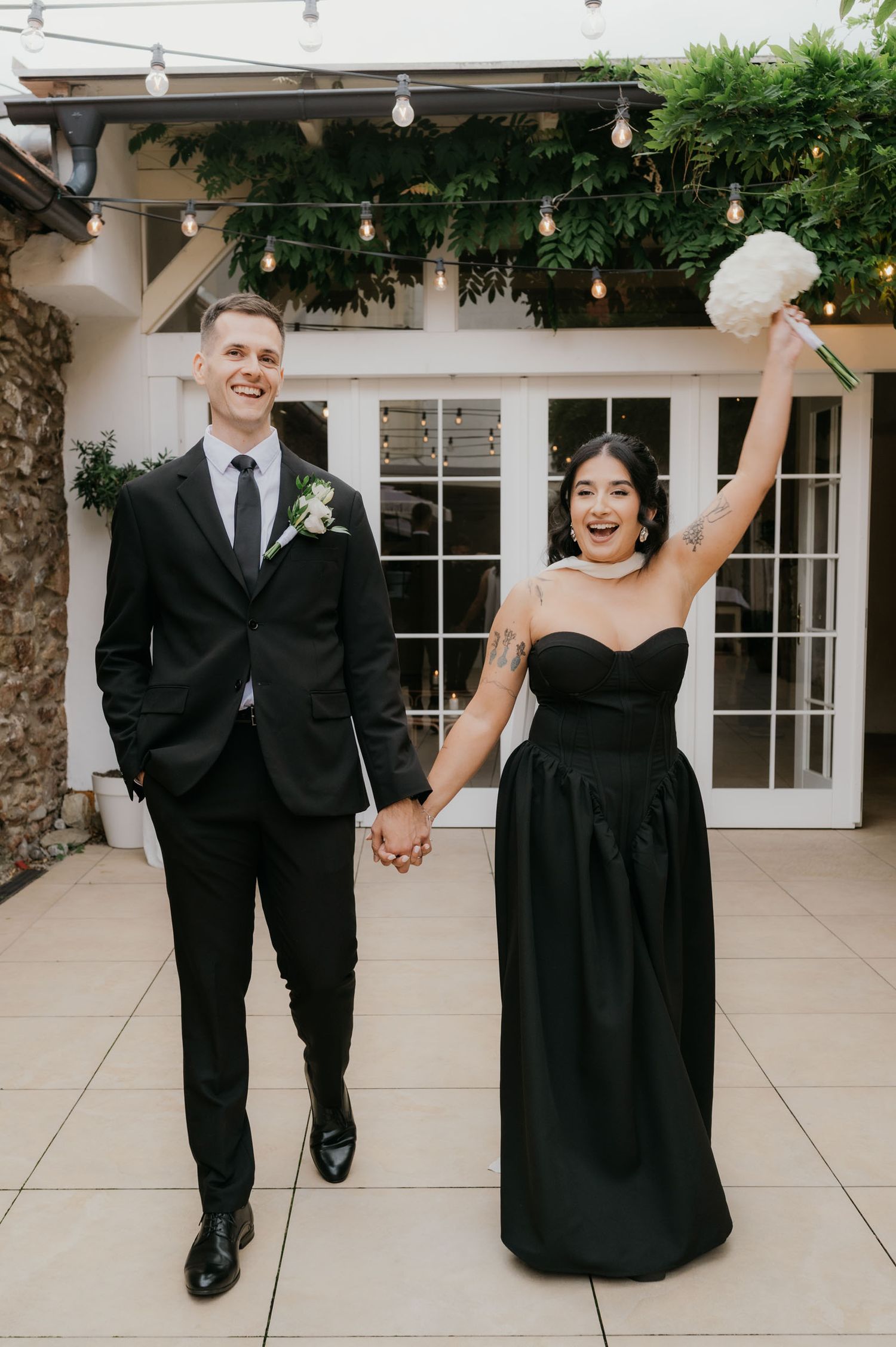 Joyful couple in matching black wedding attire holding hands, bride raising bouquet, posing outside elegant venue.