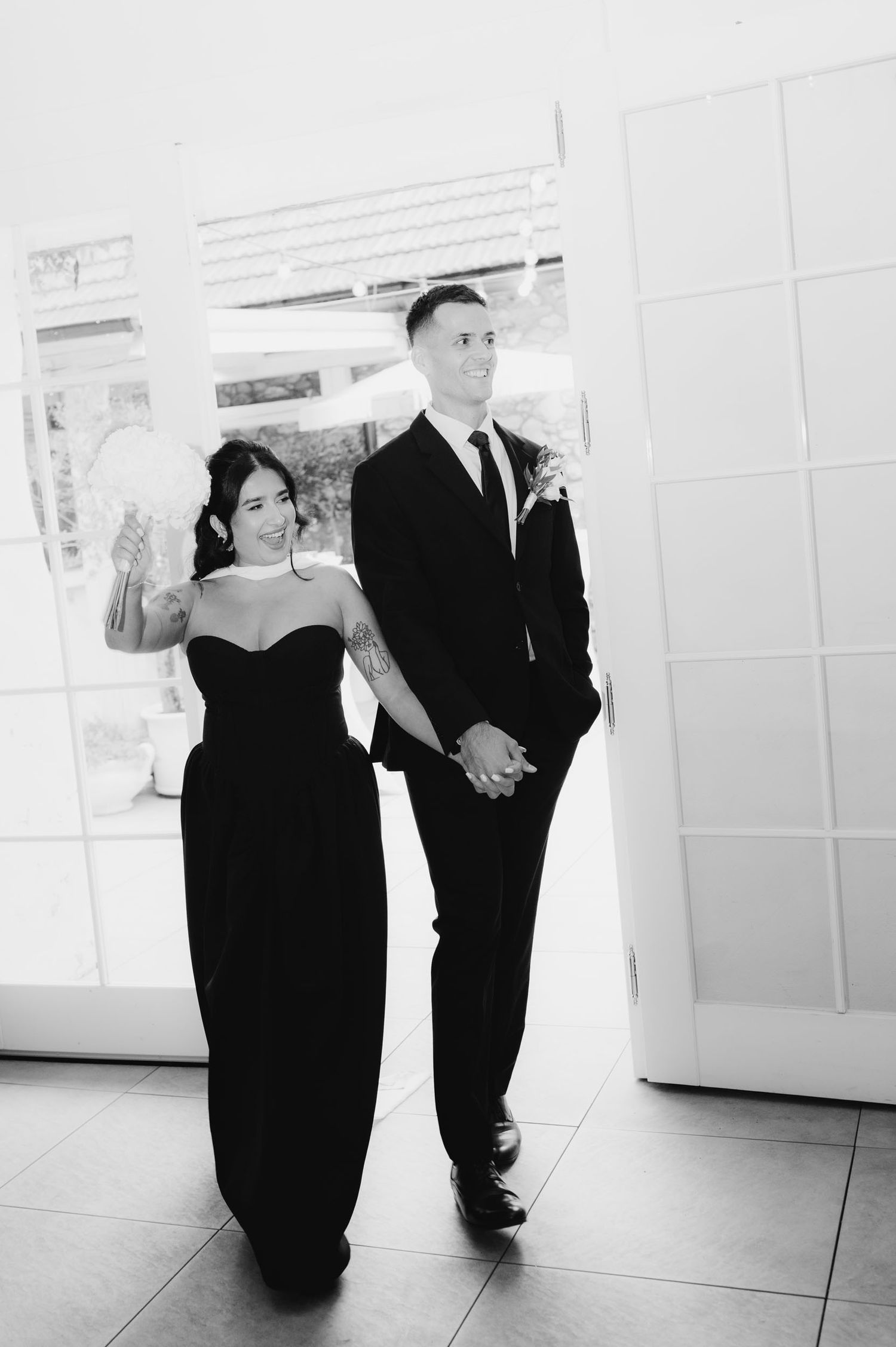 Black and white photo of a couple in formal attire, woman in strapless gown and man in suit, holding hands.