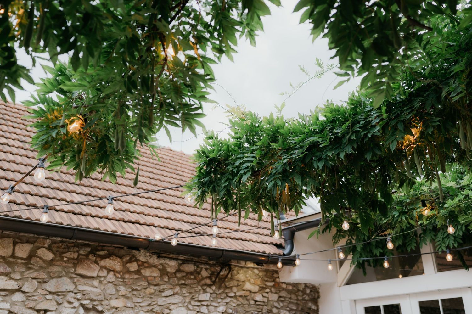 Rustic stone venue with terracotta roof, lush green garlands, and warm string lights hanging from trees.