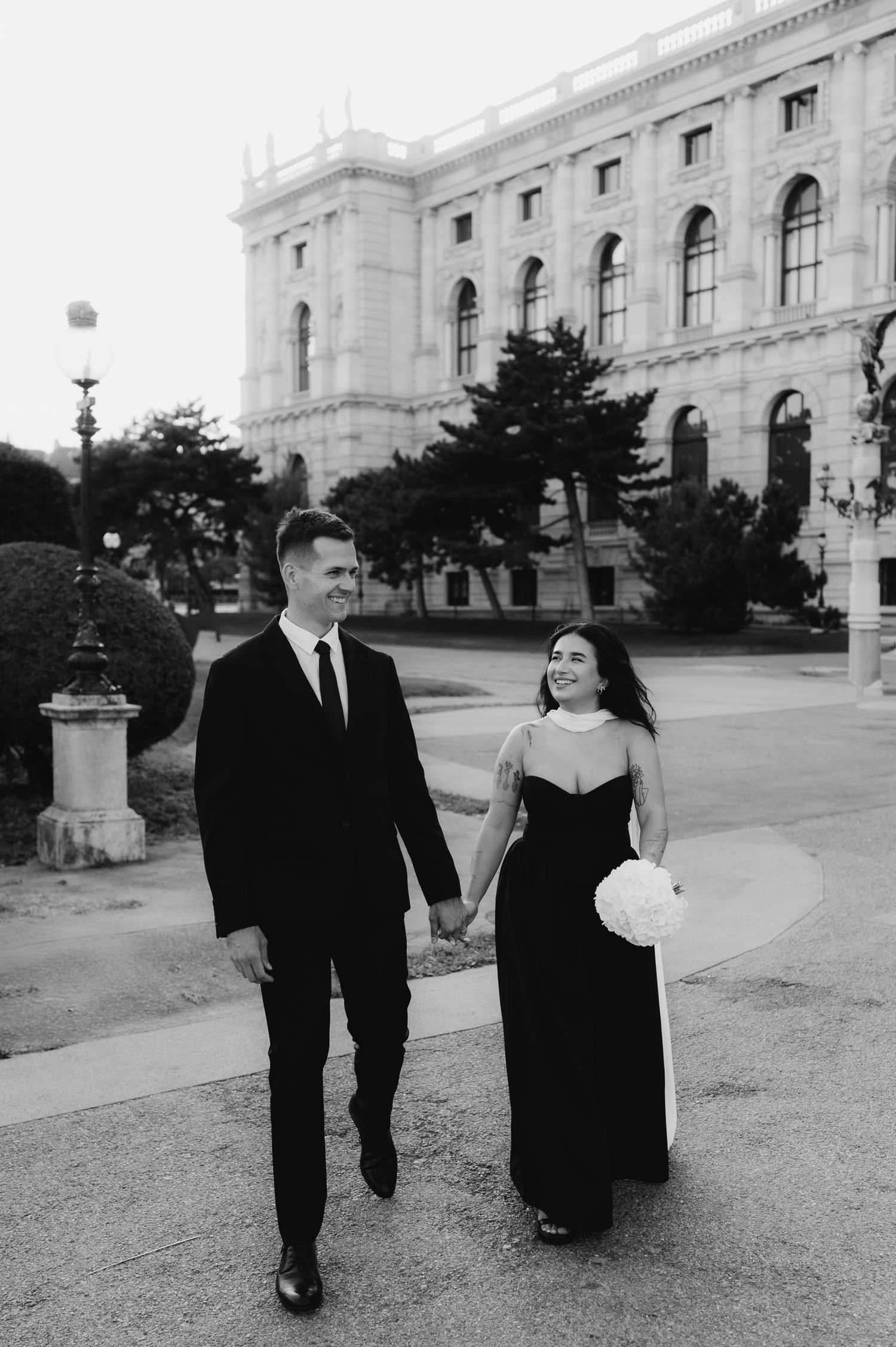 Black and white photo of a couple in formal attire holding hands and walking in front of a grand European building.