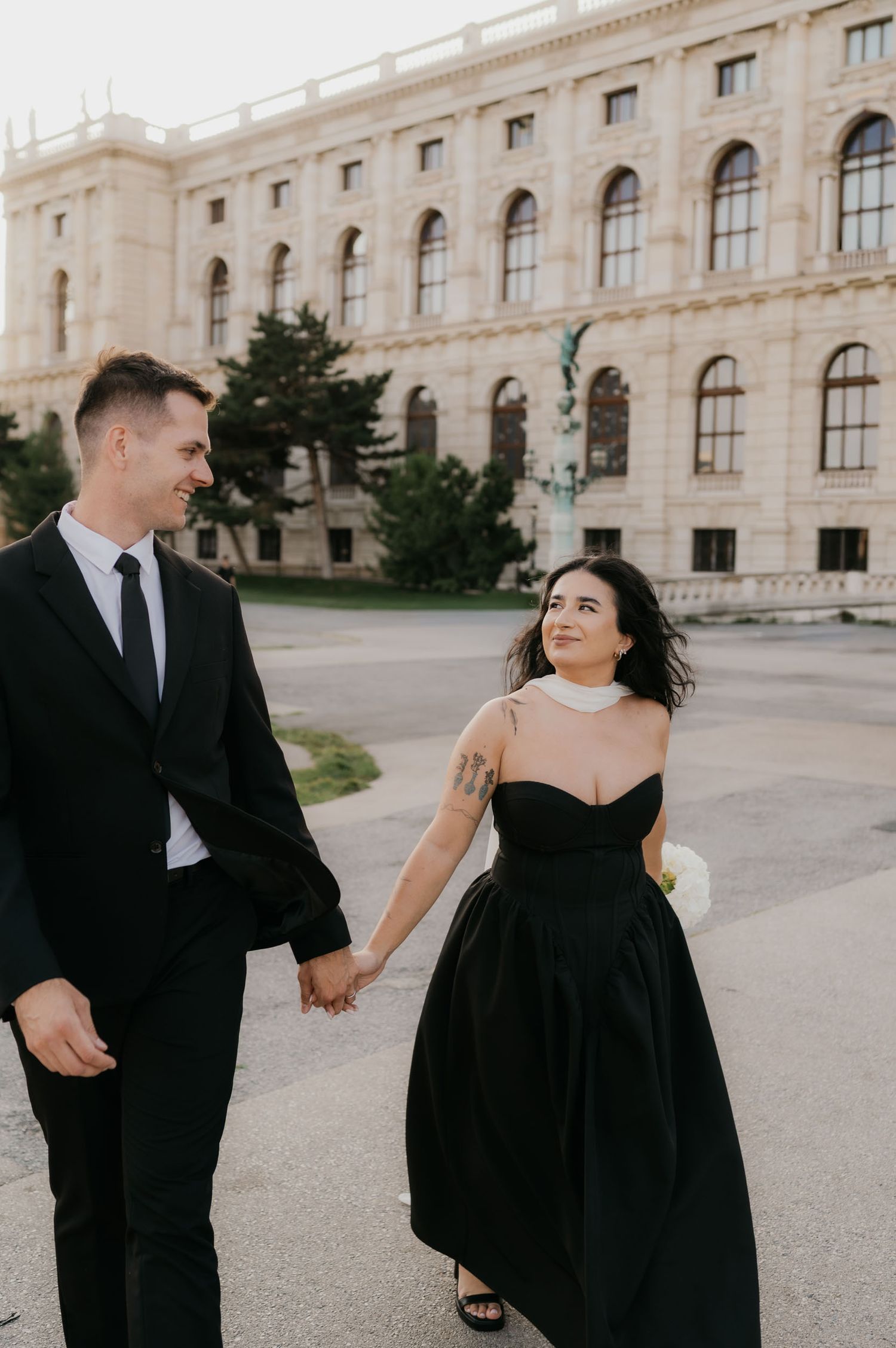 A couple in matching black formal attire holds hands and smiles while walking in front of a grand European building.
