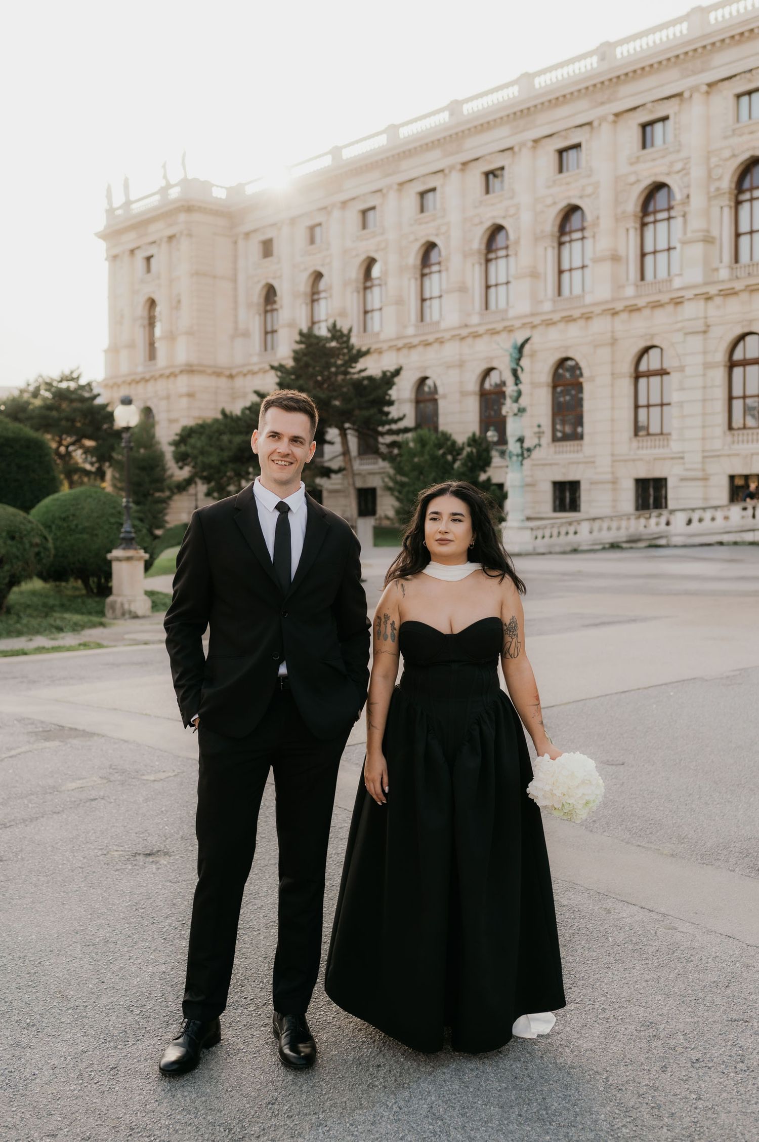Couple in matching black formal attire poses before a grand European building, woman holding white bouquet.