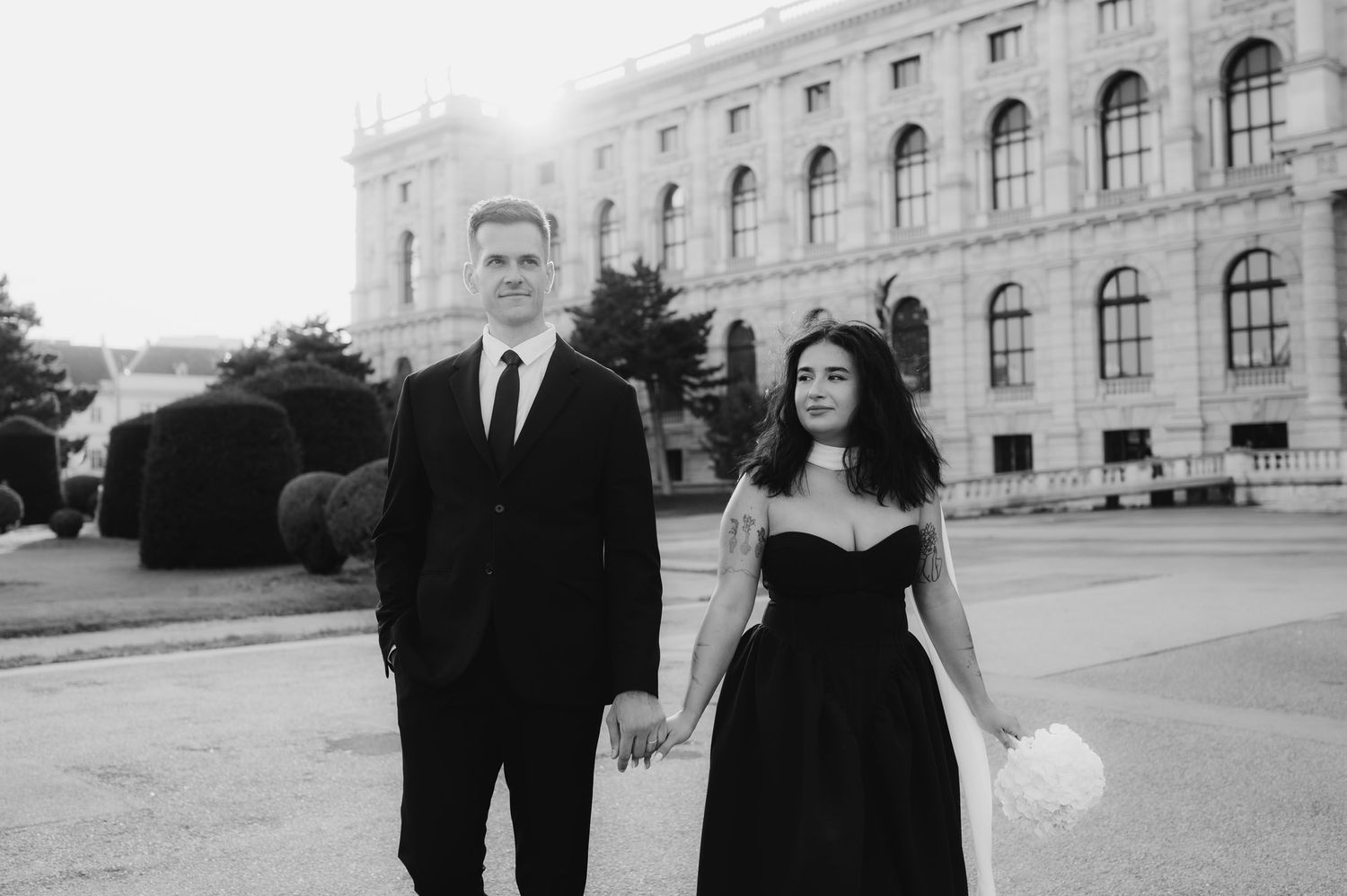 Black and white photo of an elegant couple holding hands in formal attire outside a grand historic building.