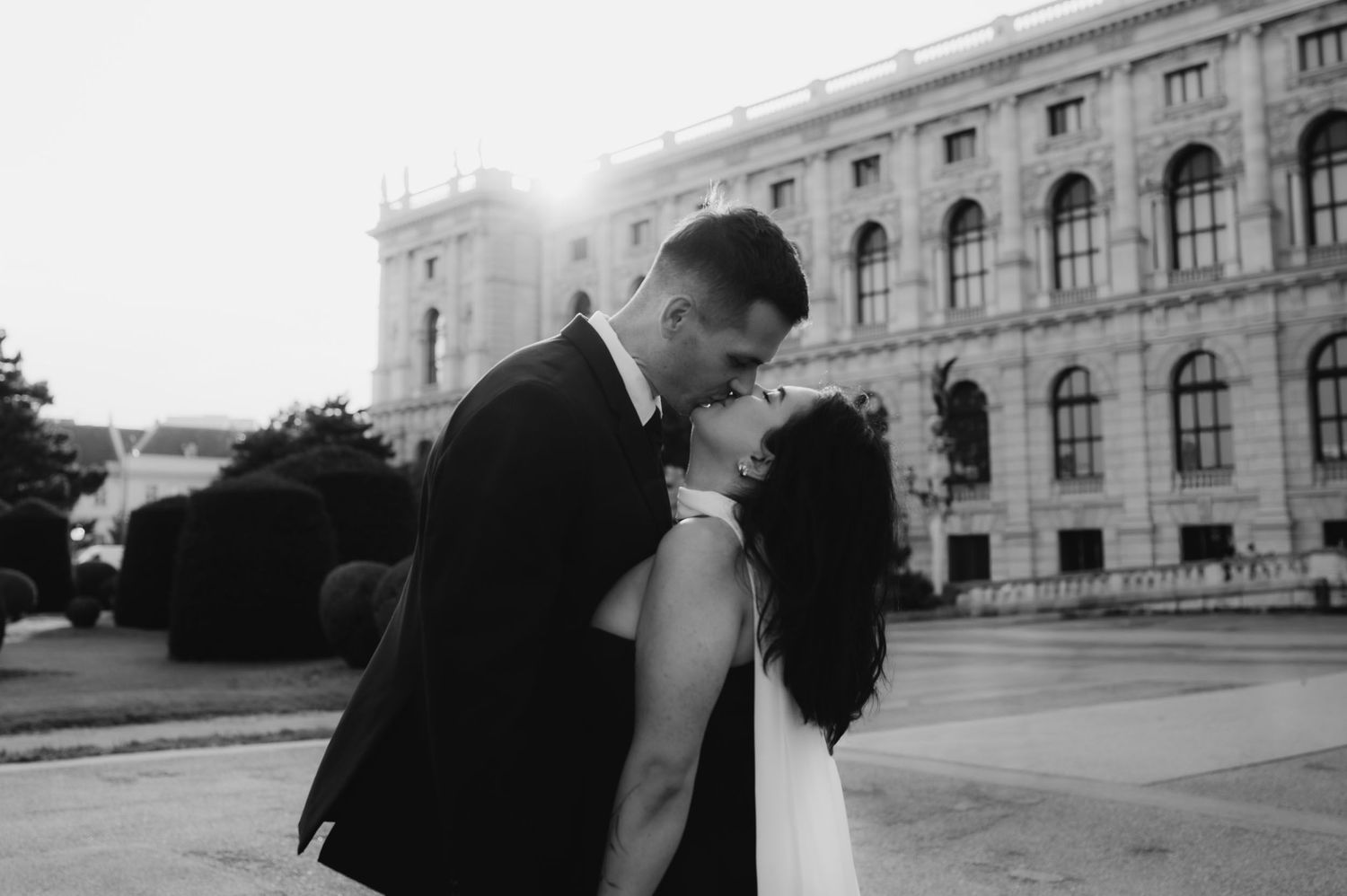 Black and white photo of a couple sharing a romantic moment near a grand European building.