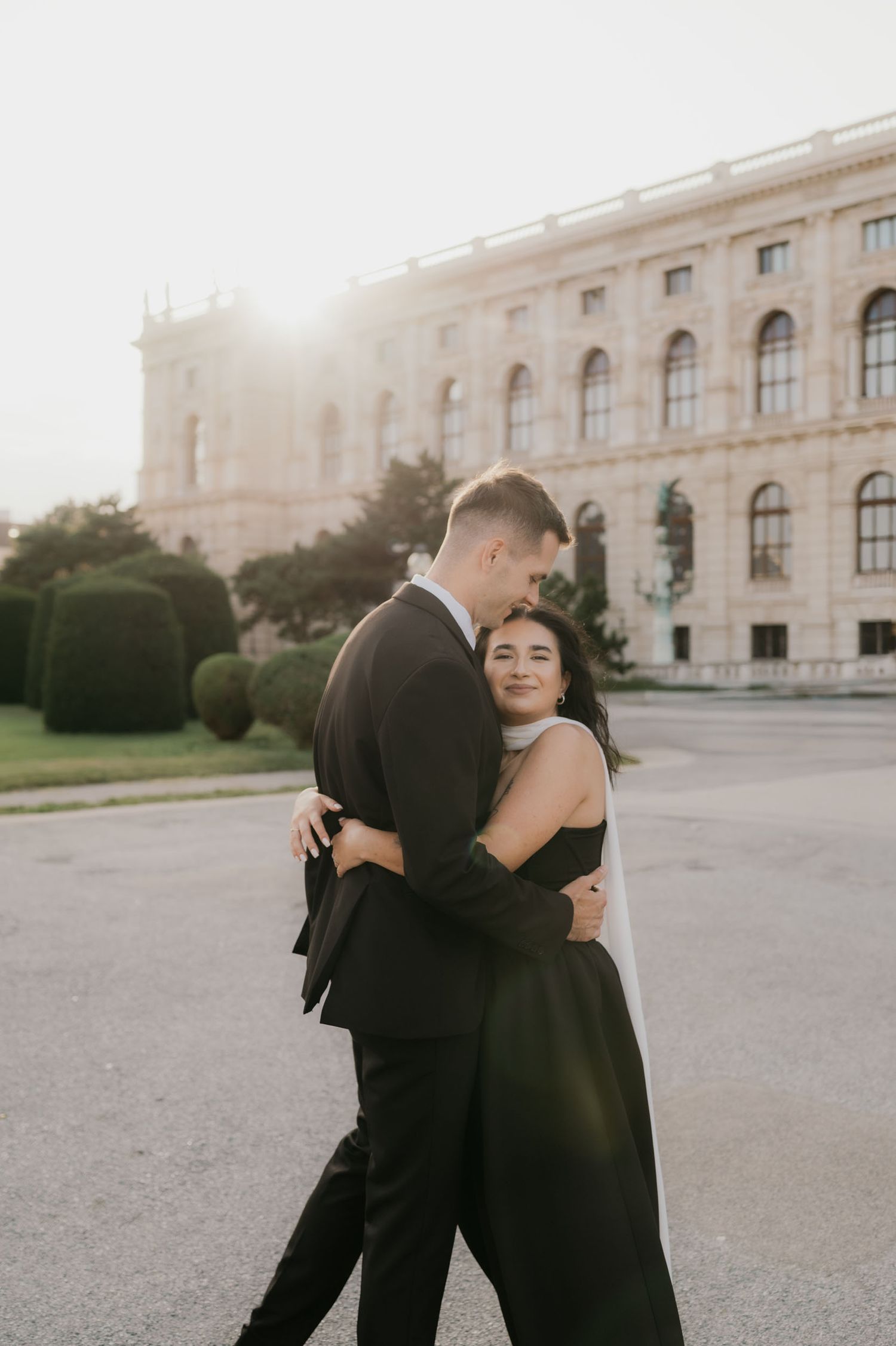 Couple embracing at sunset in front of a grand European building, dressed in elegant black formal attire.