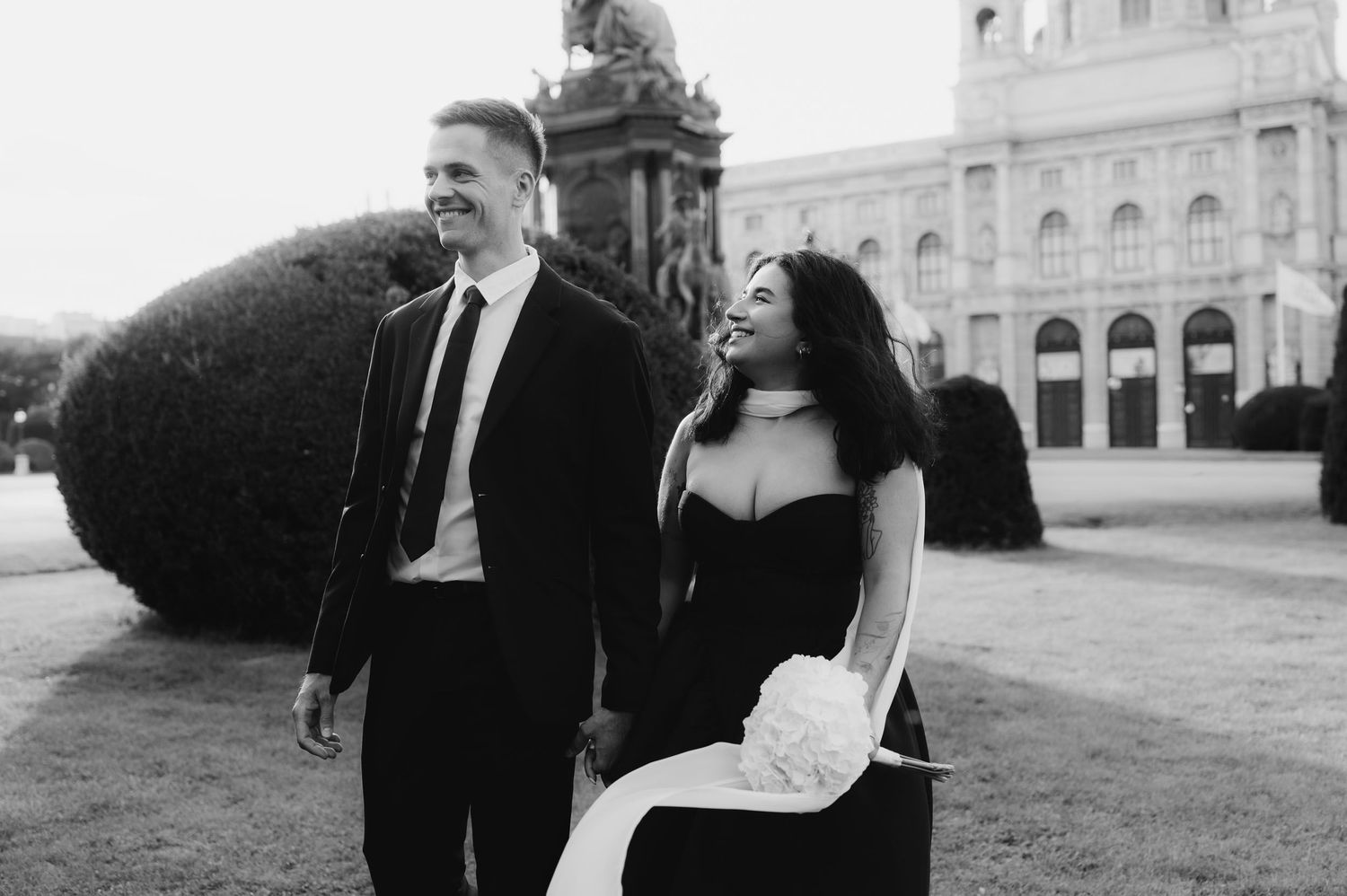 A smiling couple in formal black attire walks hand in hand outdoors, with a bouquet and elegant architecture behind them.