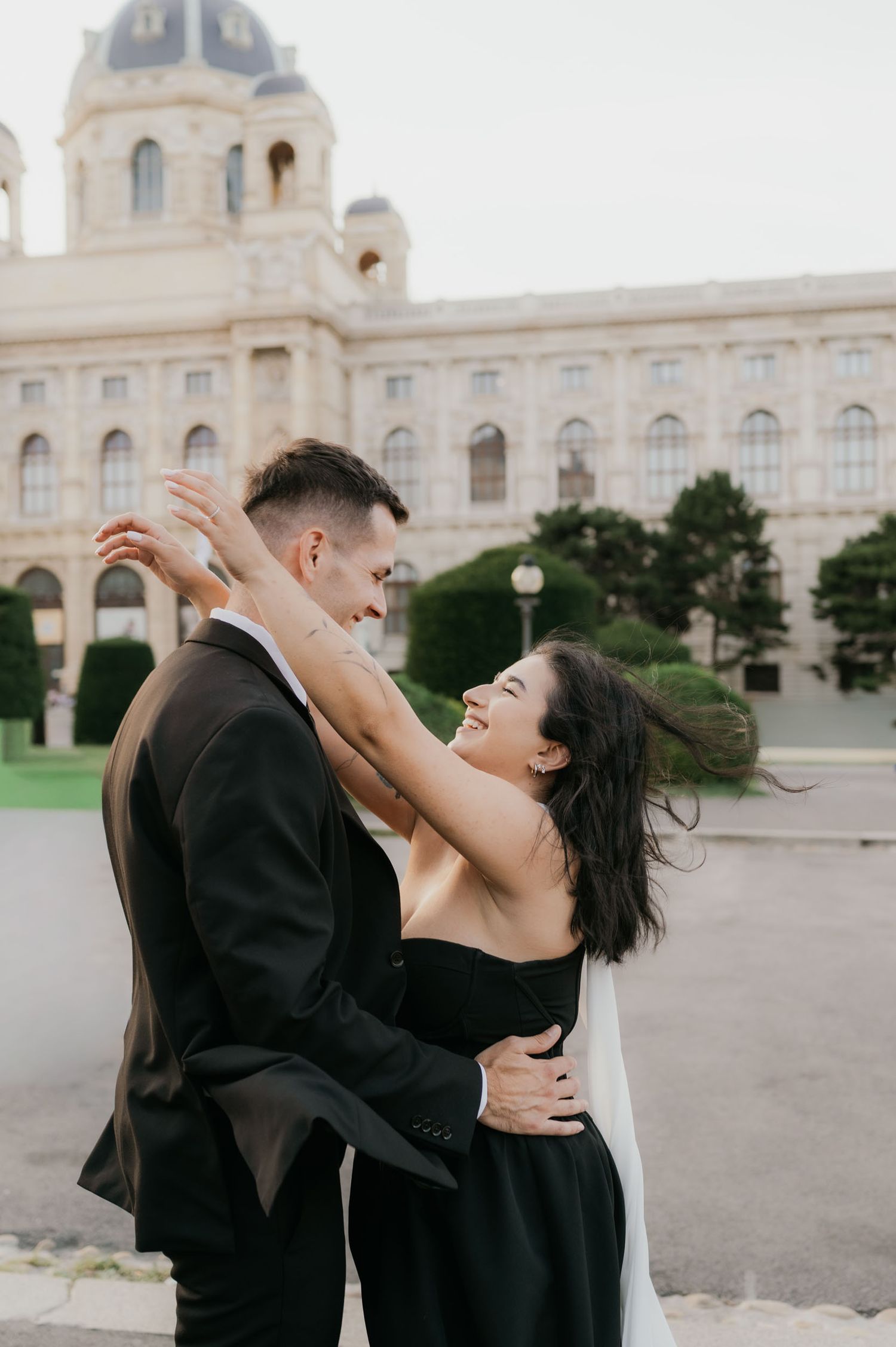 A couple in elegant black attire embraces and laughs in front of a grand European building.