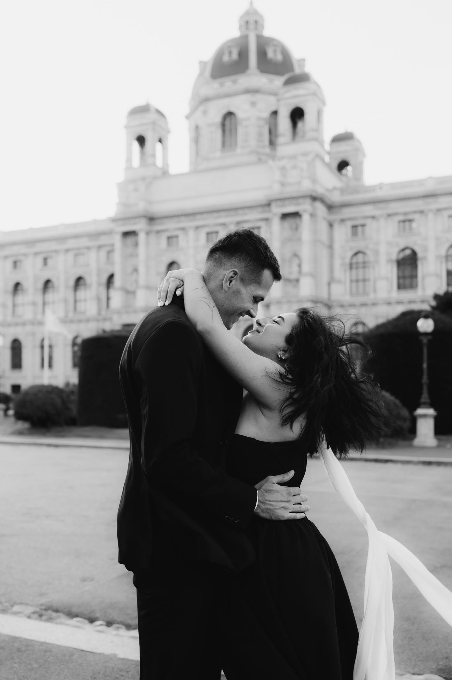 Black and white photo of a couple embracing romantically in front of a grand European building.