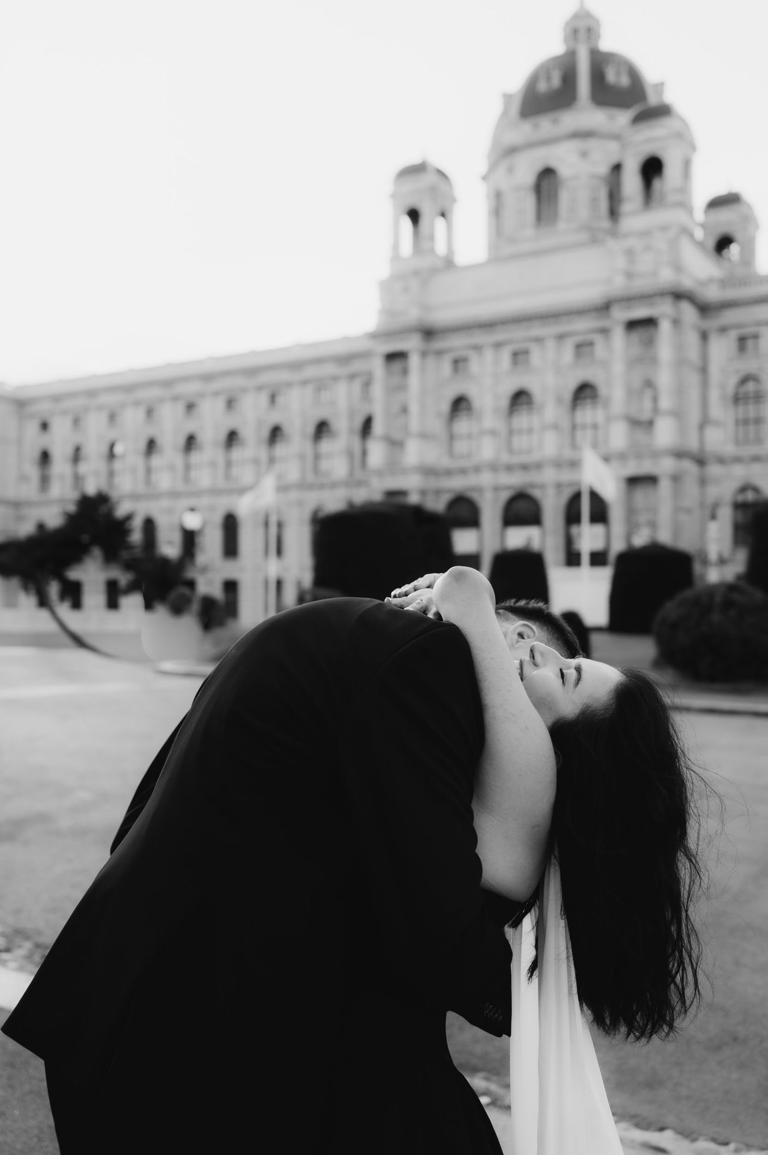 Black and white photo of a couple sharing a romantic dip kiss in front of a grand European building.