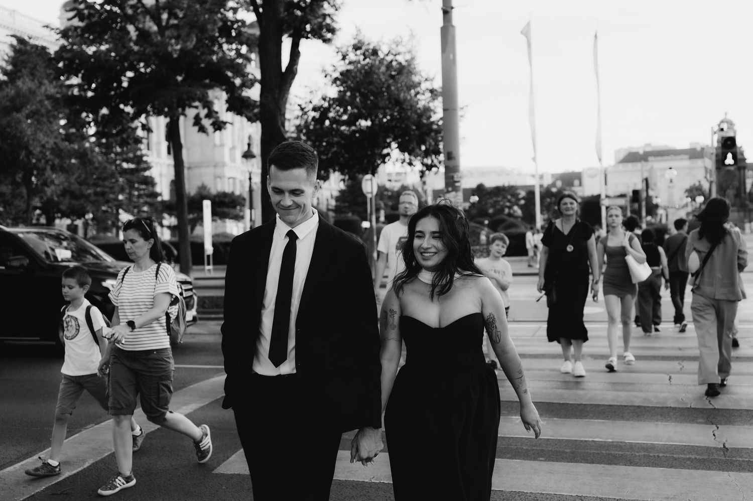 Elegant couple in formal attire holding hands while crossing a busy street in a European city.