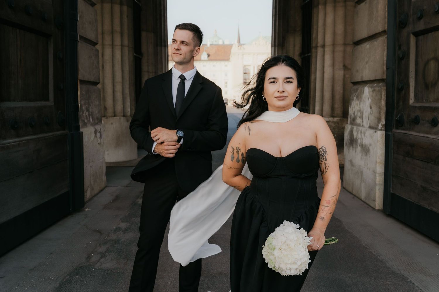 Bride in black strapless gown holding white bouquet stands before groom in black suit at elegant stone building.