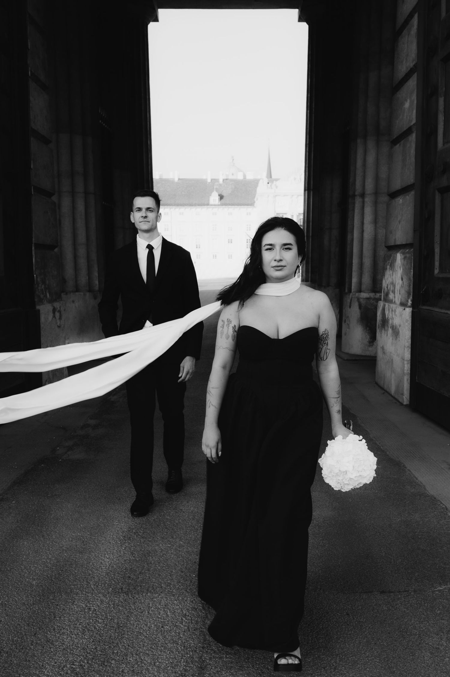 Black and white wedding photo of bride in black gown holding bouquet with groom in suit behind her.
