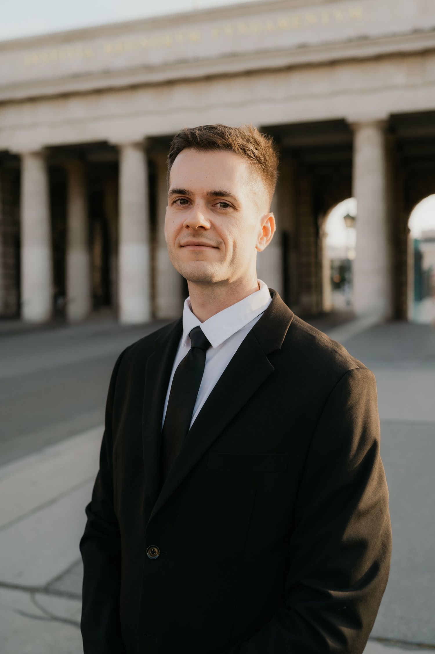 Young professional man in black suit and tie standing outdoors near classical stone columns.