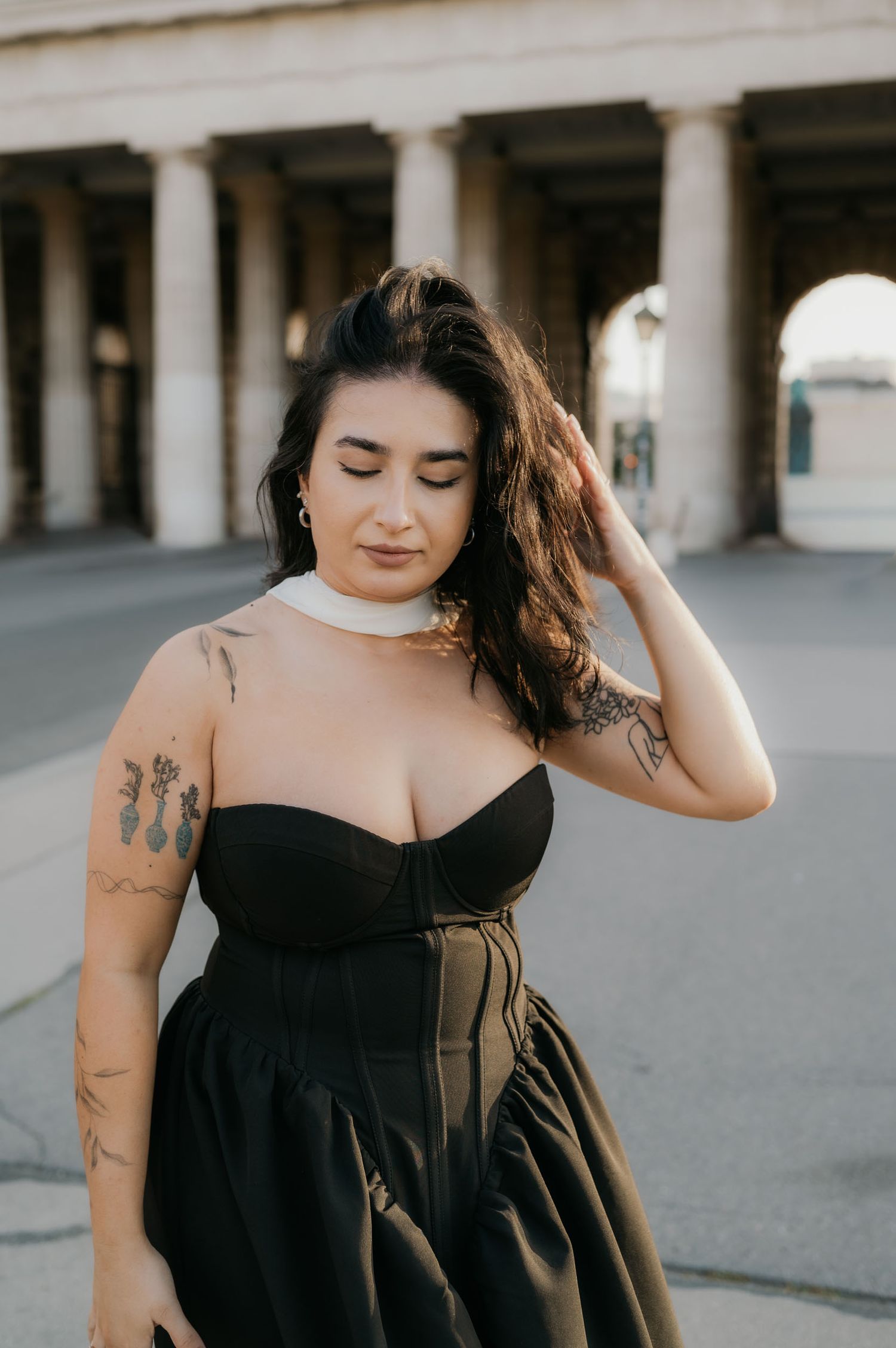Woman in black corset dress with tattoos poses outdoors near classical columns.
