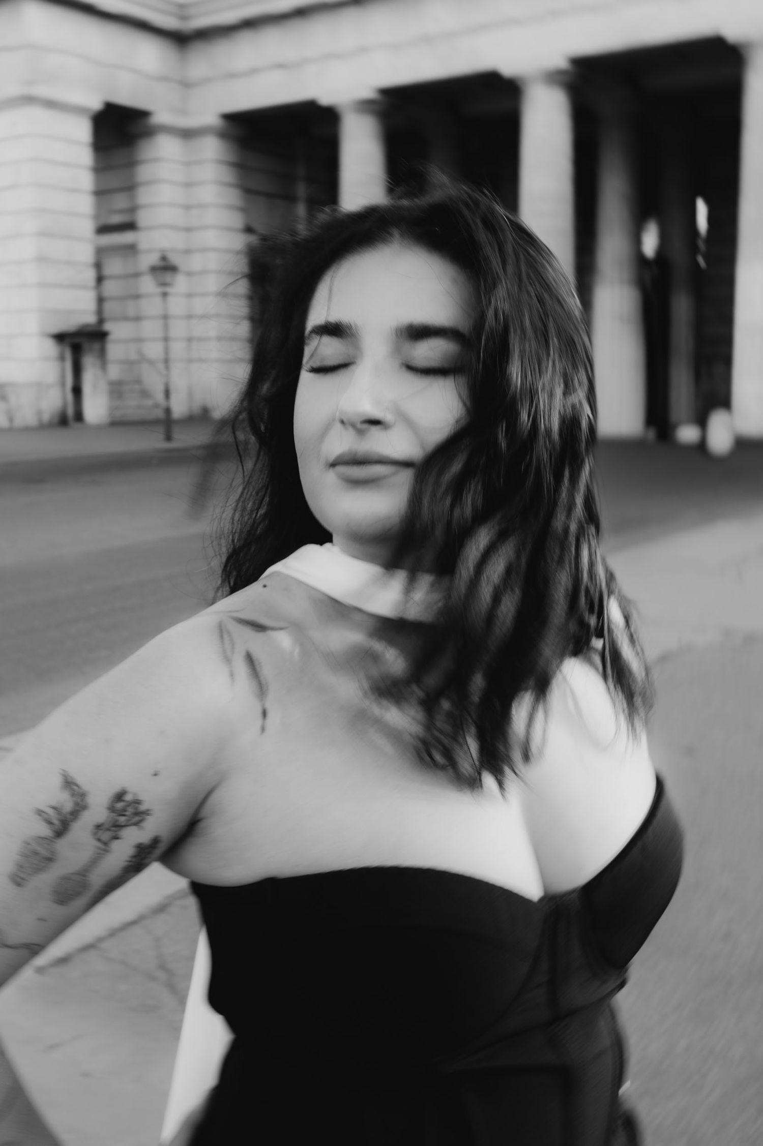 Black and white portrait of woman in black dress with tattoos, eyes closed, hair blowing in wind.