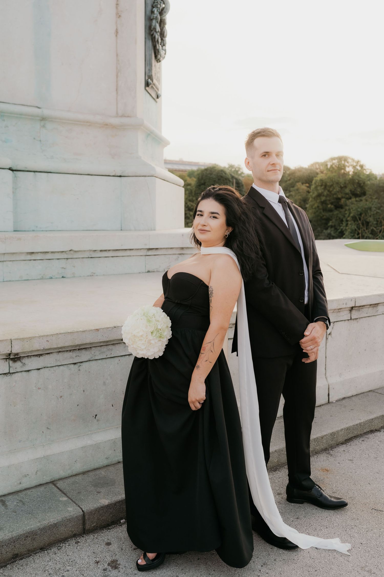 Couple in matching black formal attire pose outdoors, bride holding white hydrangea bouquet with elegant white cape train.