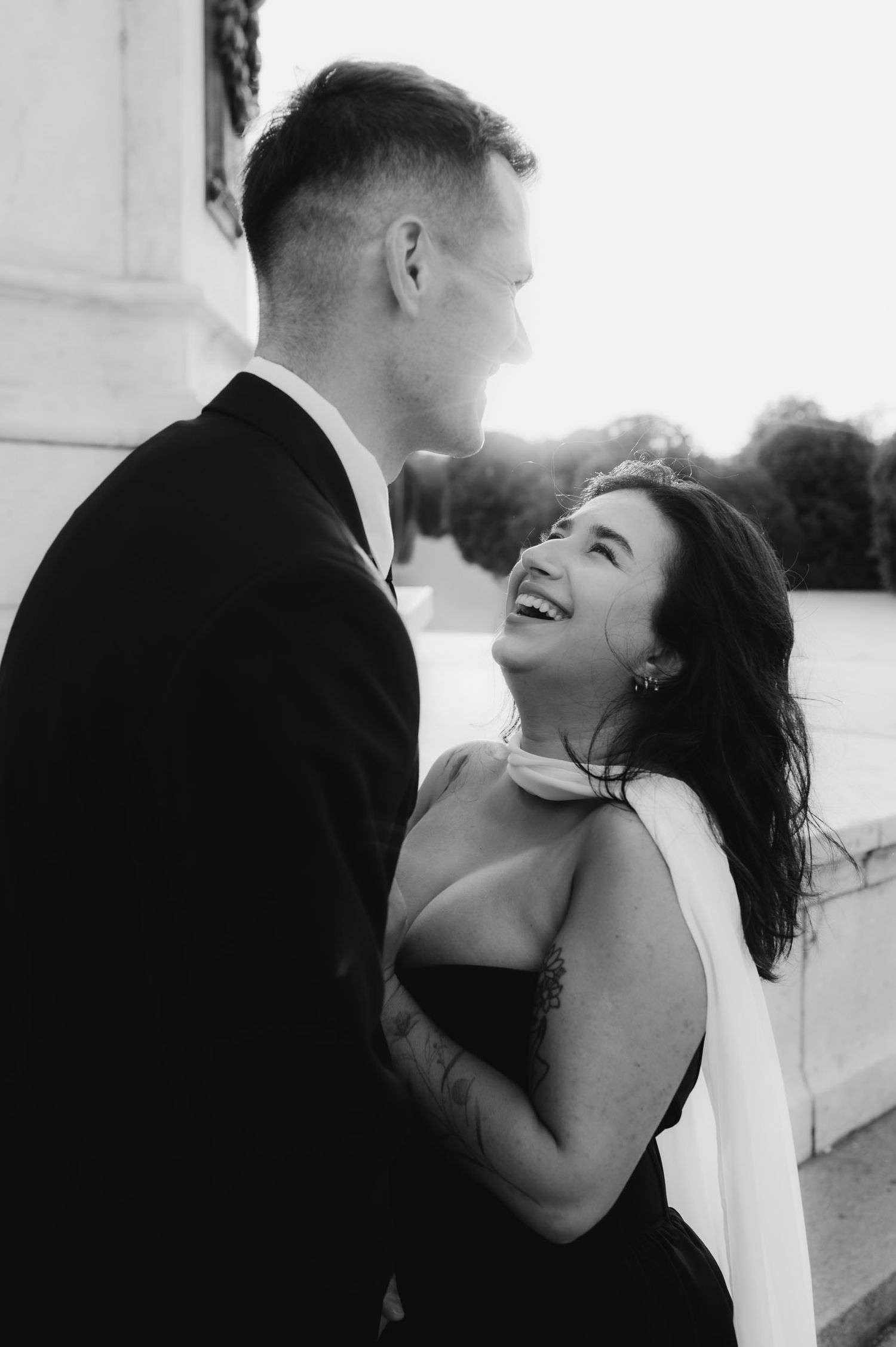 Black and white photo of laughing bride in strapless gown gazing up at groom in black suit outdoors.