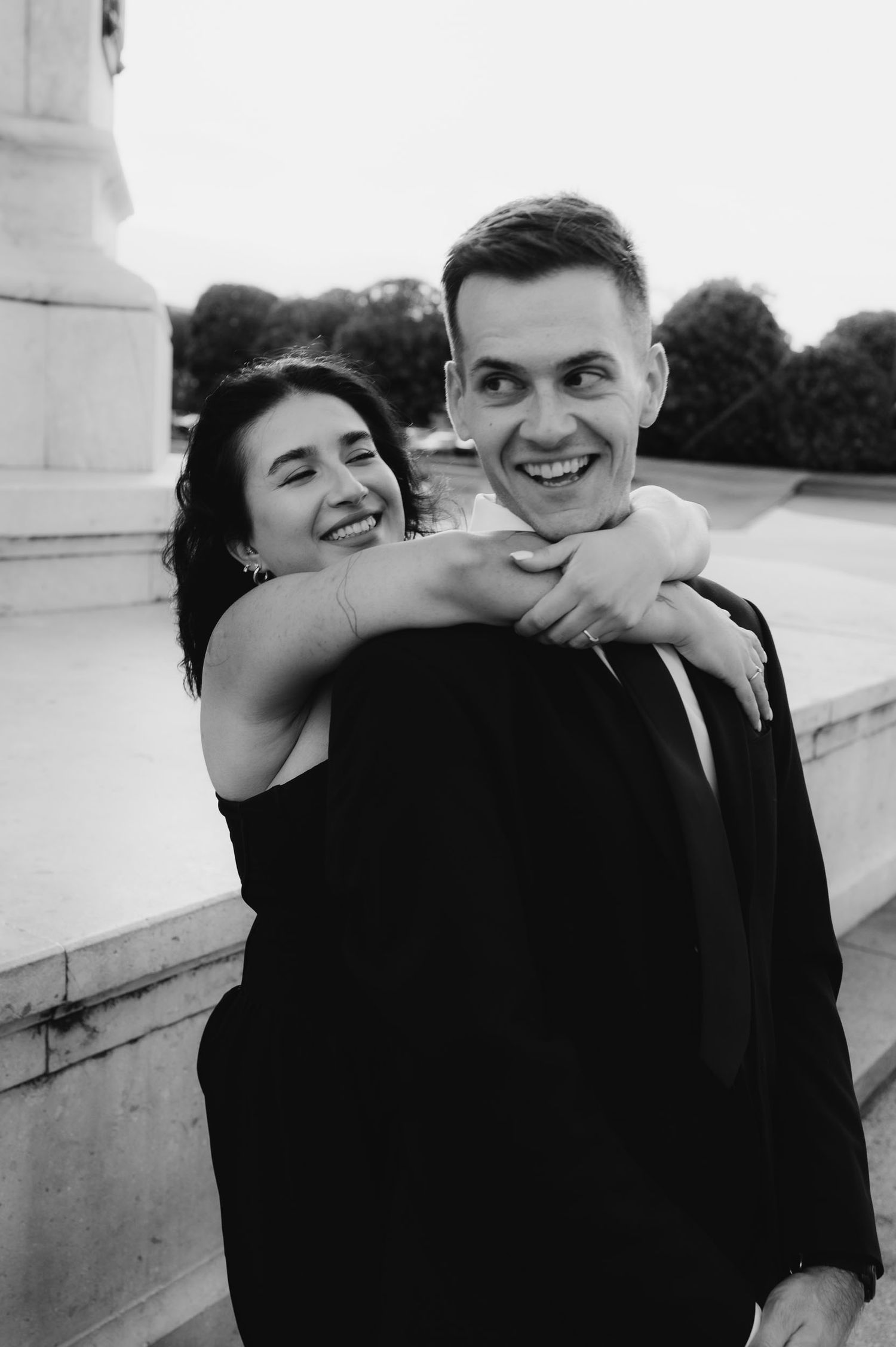 Black and white photo of a smiling couple, woman hugging man from behind, both dressed in elegant attire outdoors.