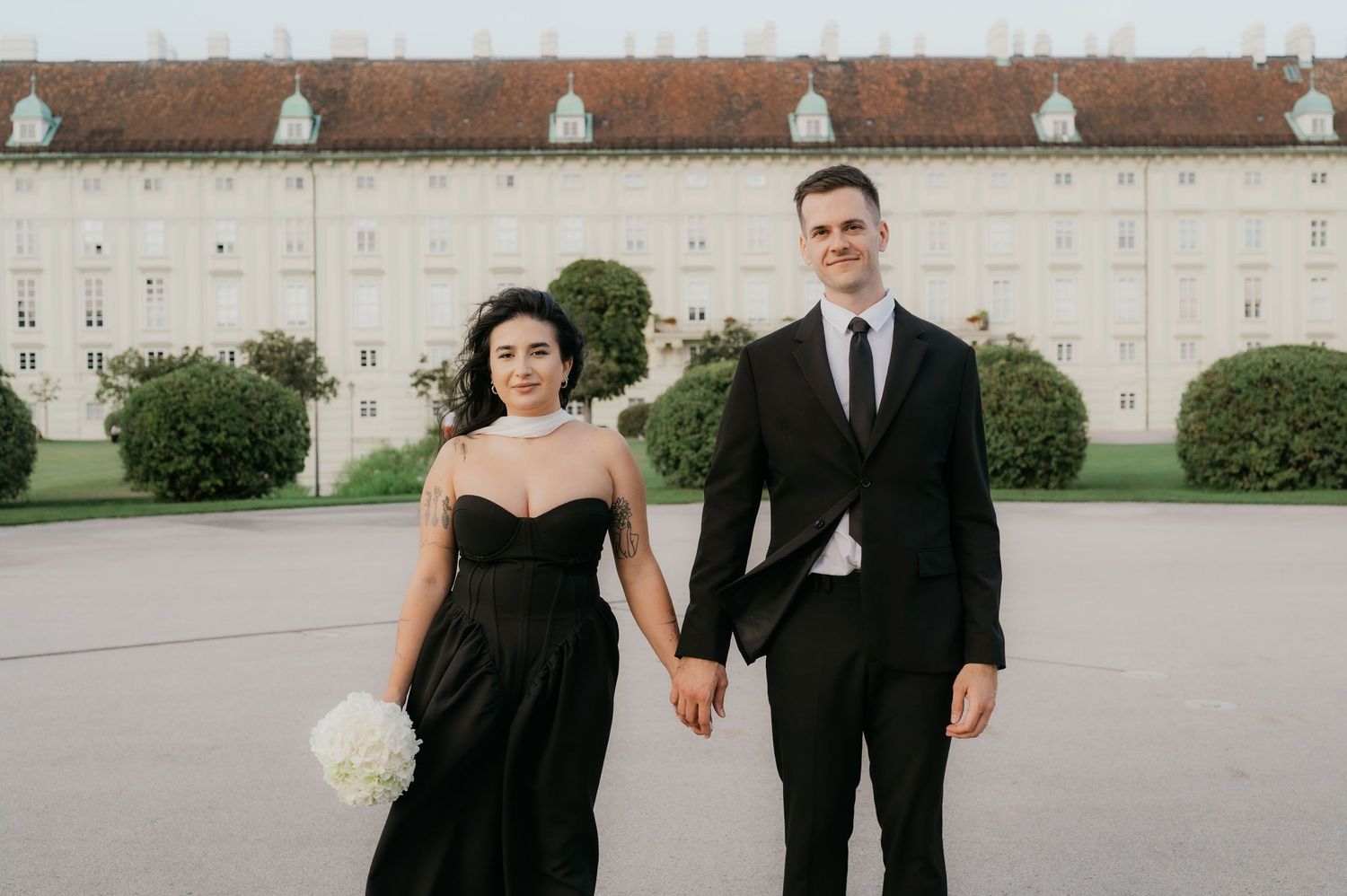 Couple in matching black formal attire holding hands before a grand white European building.