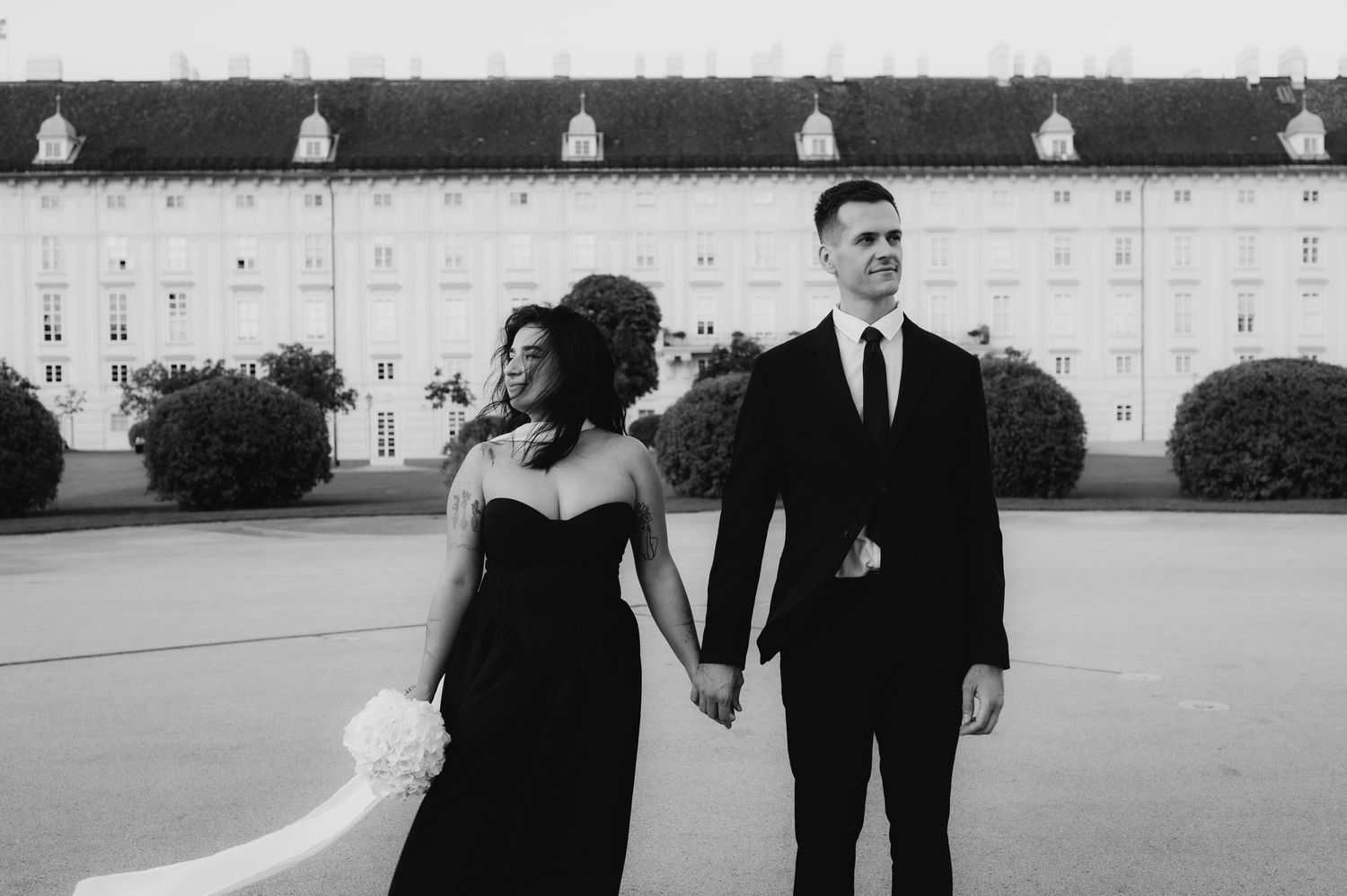 Black and white photo of couple in black formalwear holding hands outside elegant building.