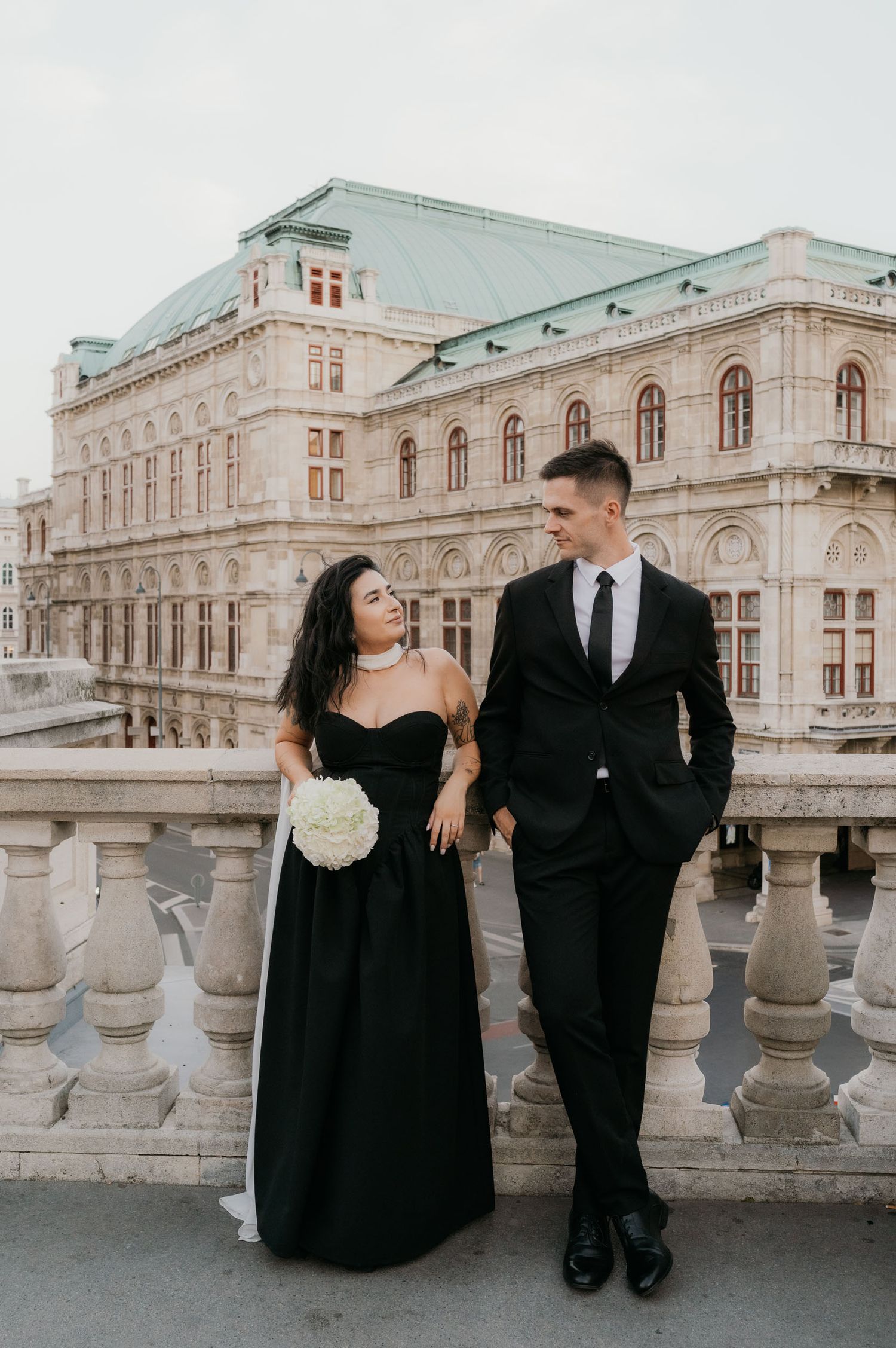 Couple in matching black formal attire poses on ornate balcony before grand European building with green copper roof.