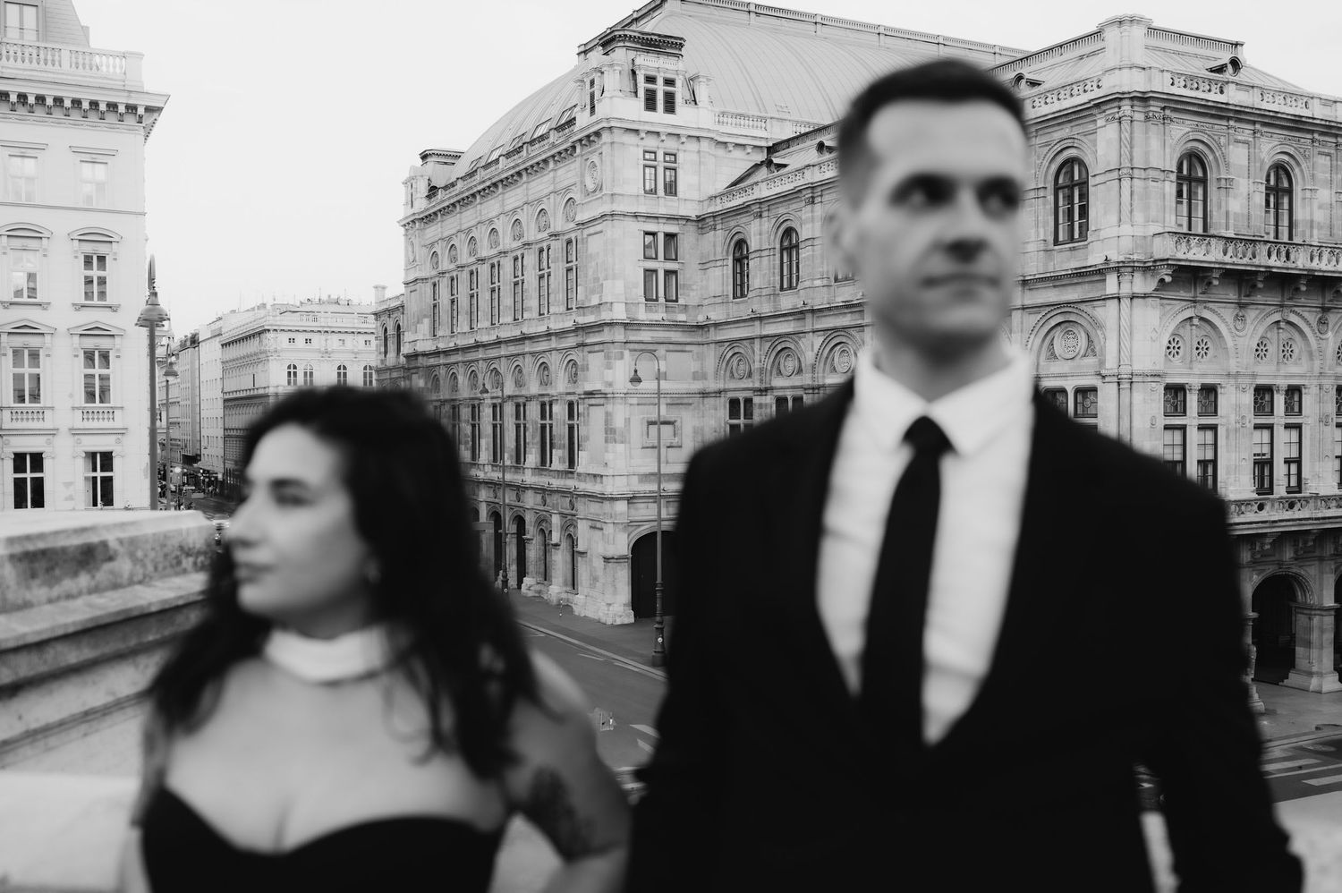 Black and white photo of an elegant couple in formal attire walking near a grand European opera house.