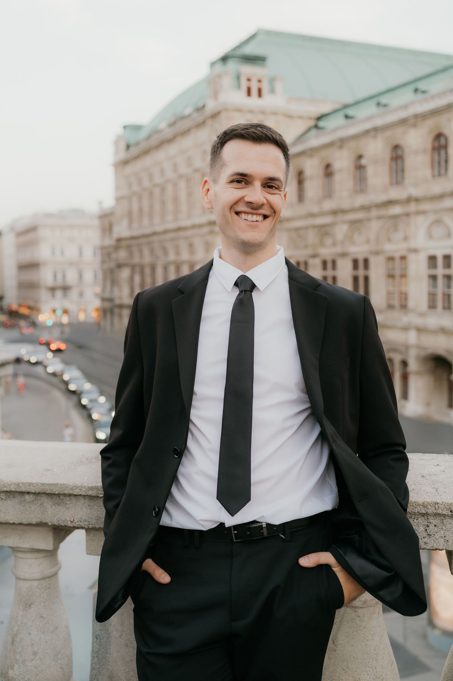 Smiling young man in black suit and tie posing outdoors with historic European building in background.