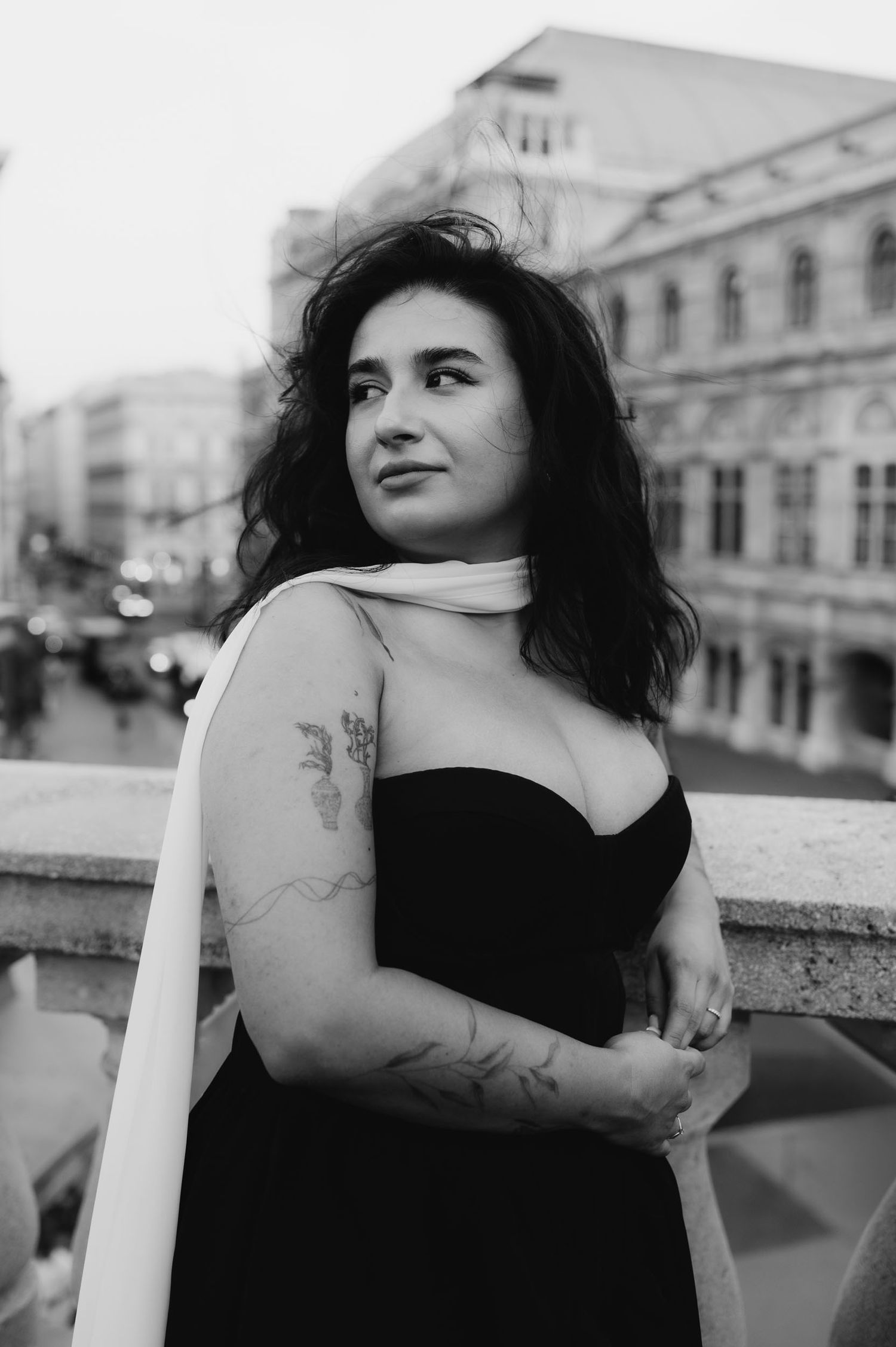Black and white portrait of a tattooed woman in a strapless dress with a white shawl, posing on a bridge in a European city.