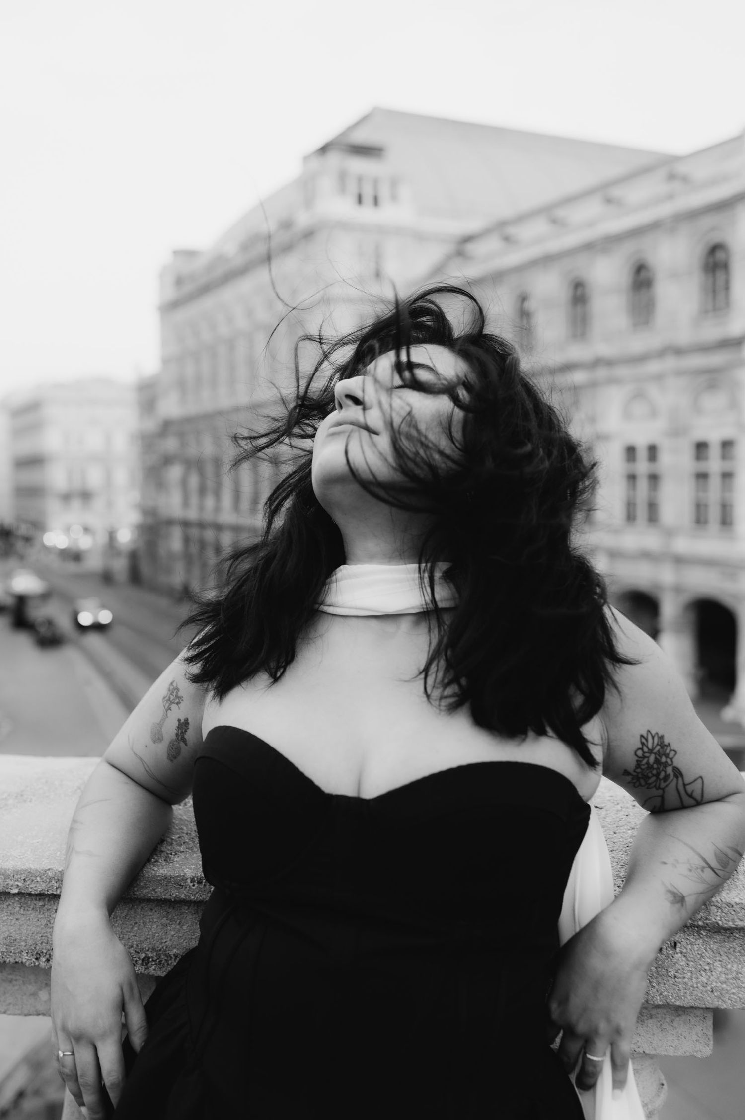 Black and white photo of a woman with wind-blown hair in a black strapless dress and choker, posing on a balcony.
