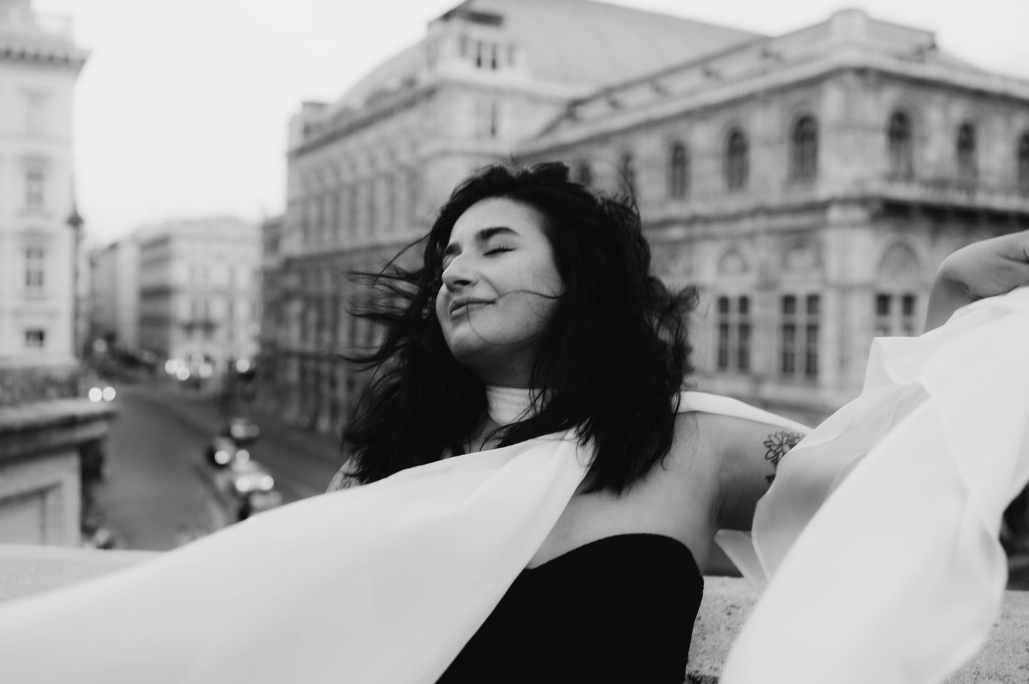Black and white photo of a joyful woman with flowing hair and fabric, posing on a balcony with a European cityscape.